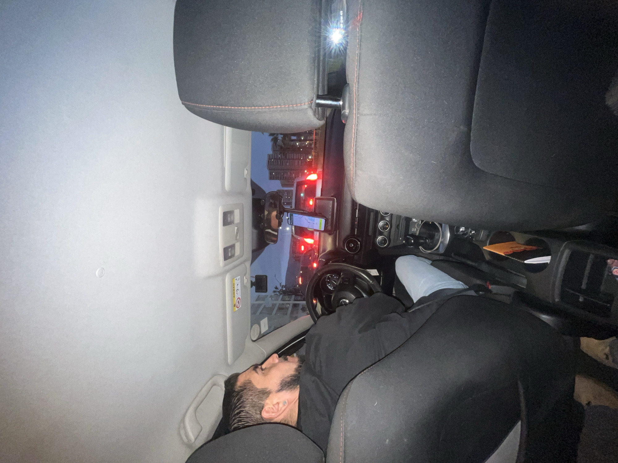 The image shows the interior of a vehicle, likely a car, from a rear-facing perspective. A man with a beard is driving, looking to his left. He is wearing a black sweatshirt and blue jeans. The car's dashboard and steering wheel are visible, along with a mounted phone displaying a navigation app. Outside the car, a cityscape with buildings and streetlights can be seen, suggesting an urban environment at dusk or night. The car appears to be on a road, possibly in Iquique, Chile, given the architecture visible in the background, which resembles buildings commonly found in coastal cities of South America. The seats and interior of the car are dark-colored. The scene conveys a sense of travel or transit within a city.