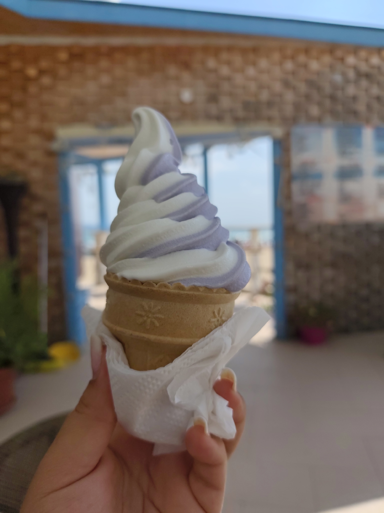 A hand holds a swirled soft-serve ice cream cone, featuring alternating bands of white and light purple, wrapped in a white napkin. The cone itself is a light tan waffle cone with a embossed star pattern around its rim. In the background, a rustic brick building with blue-trimmed windows and doorways suggests a setting in Nesebar, Bulgaria. Out of focus, glimpses of greenery and possibly the sea can be seen through the doorways, hinting at an outdoor or semi-outdoor establishment. The lighting suggests it is daytime.