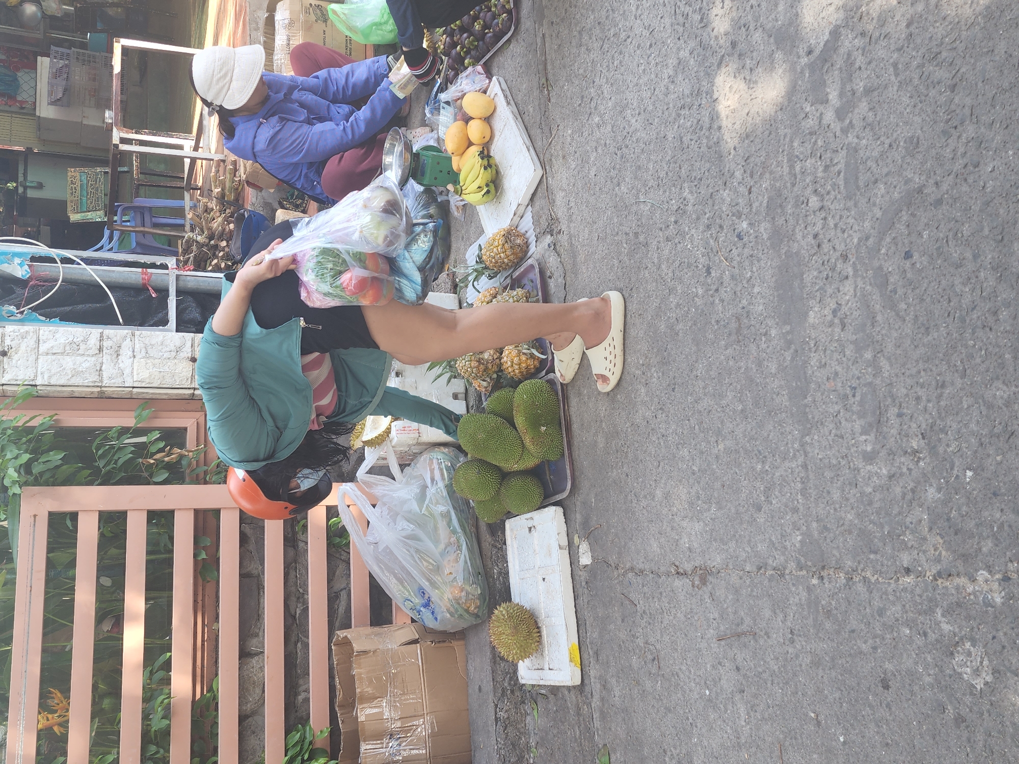 The image depicts an outdoor market scene, likely on a street in Phu Thuy Ward, Vietnam, given the presence of tropical fruits. A woman wearing a green jacket, black shorts, and an orange helmet is bent over, holding a plastic bag filled with produce, including tomatoes and leafy greens. She is standing on the right side of the frame, with her back mostly to the camera. To her left, a second woman, wearing a blue jacket and maroon pants, is seated on the ground and appears to be examining some items, possibly money or receipts. She has a white hat on. In the foreground and midground, various fruits are displayed on the ground, including durians, pineapples, bananas, mangoes, and mangosteens. Some are on white styrofoam trays, while others are loose on a black tray. Cardboard boxes are visible in the background, along with a metal fence and some greenery. The ground is paved with concrete. The scene suggests a commercial transaction or preparation for sales at a street-side market.