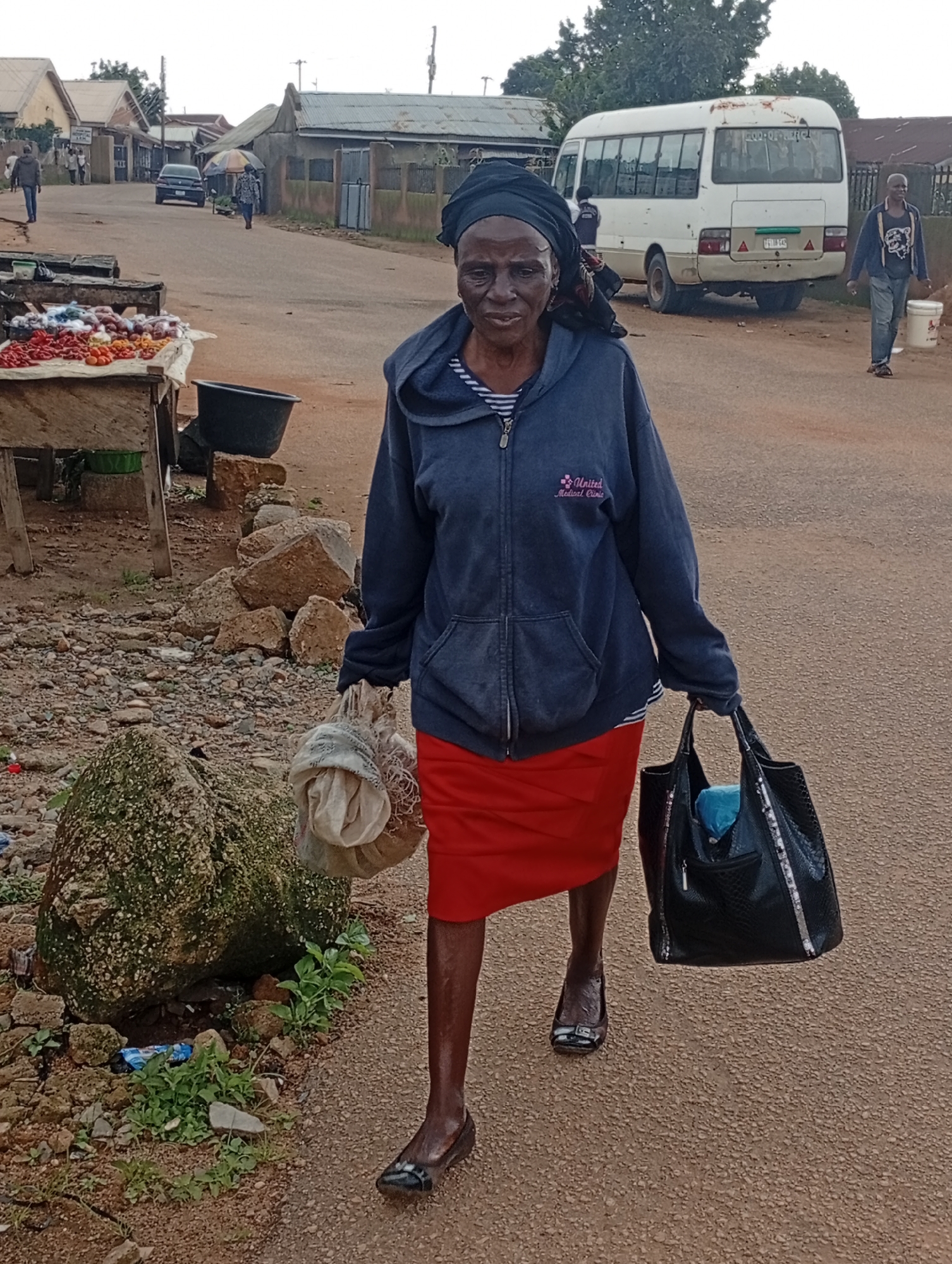 The image depicts a street scene in Jos, Nigeria, with an elderly woman walking towards the viewer. She is wearing a dark blue hooded jacket over a striped shirt and a red skirt. In her right hand, she carries a bundle wrapped in light-colored fabric, and in her left hand, she holds a black patterned tote bag. The street appears to be unpaved, with a rough texture. To the left of the woman, a market stall is visible, laden with red and yellow produce. Rocks and vegetation line the side of the road. In the background, several buildings with corrugated metal roofs are present, and a white minibus is parked on the street. Other individuals and vehicles are also present in the distance, suggesting a typical day in a Nigerian urban environment.