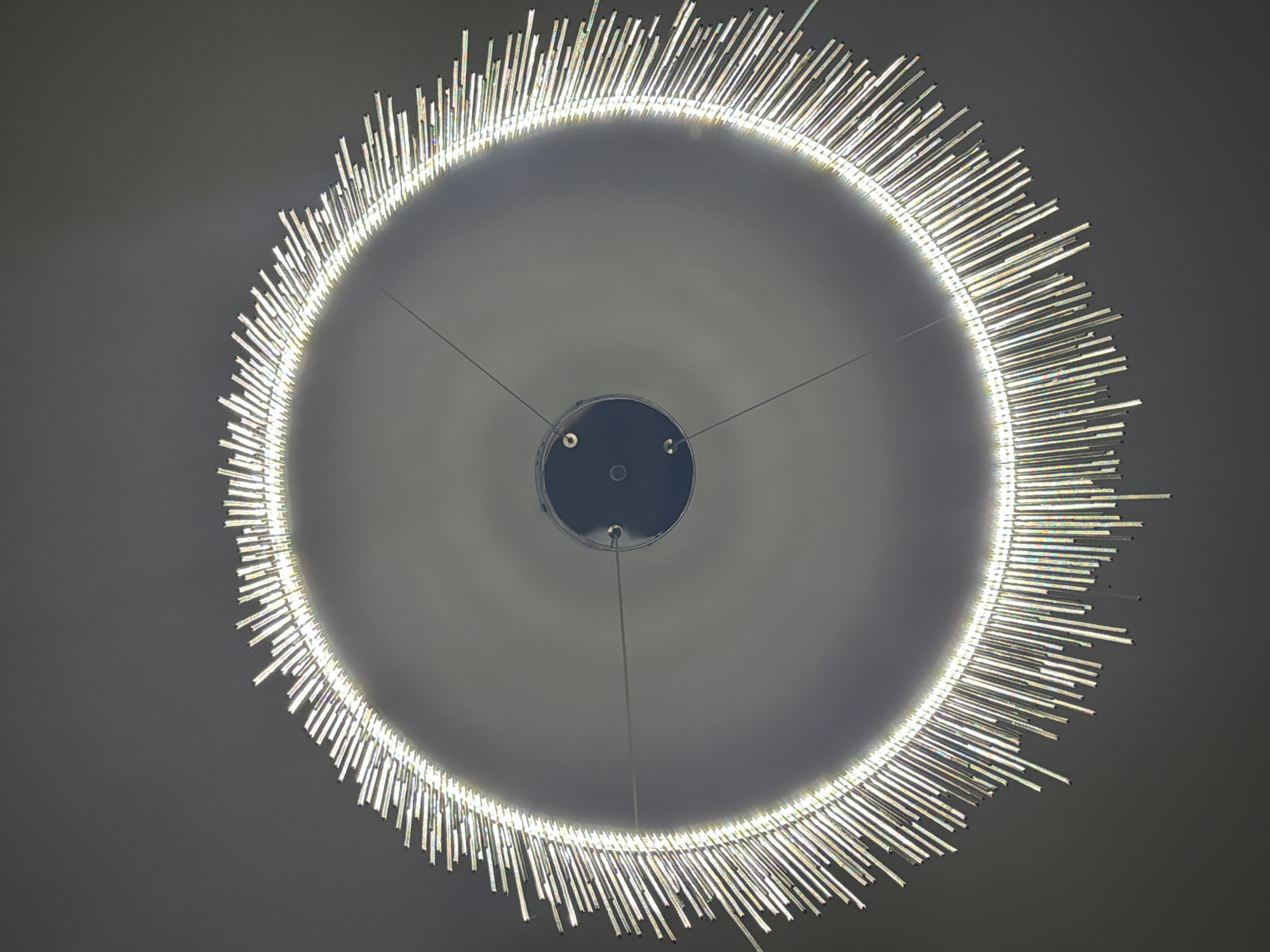 The image shows a close-up, downward view of a modern, circular chandelier hanging from a ceiling. The chandelier is composed of numerous clear, rod-shaped elements arranged in a radiating pattern around a central dark, circular fixture. These elements appear to be made of glass or crystal, as they reflect light and exhibit prismatic colors. The light source is integrated into the inner ring of the chandelier, creating a bright halo effect and illuminating the hanging rods, which cast subtle shadows on the plain, grey ceiling. Three thin wires are visible, suspending the central fixture from the ceiling. The overall impression is one of contemporary elegance and subtle illumination. This image does not depict any specific location in the city of Dubovaya, Russia, nor does it contain any people, actions, or interactions.