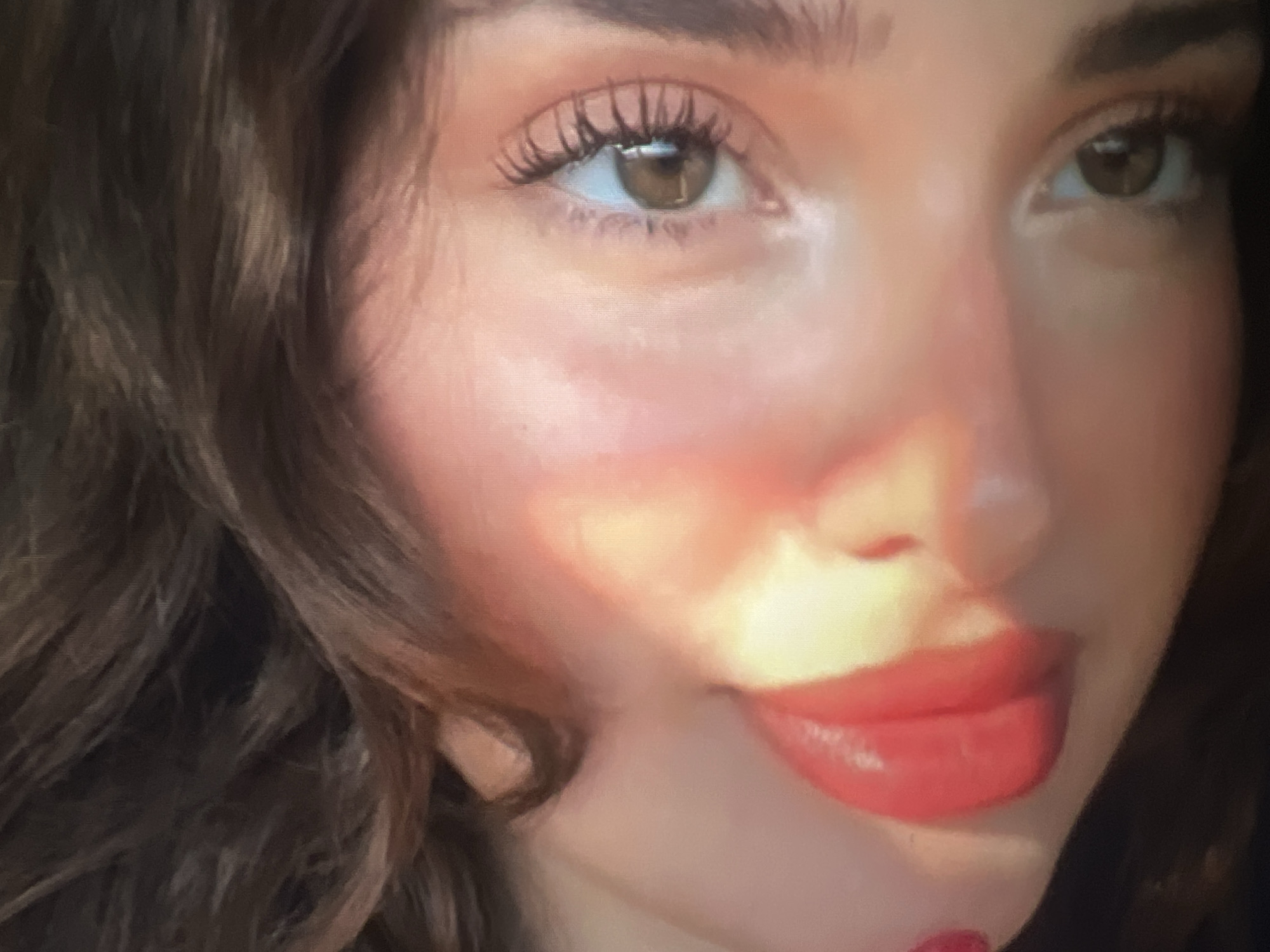 The image is a close-up, tilted portrait of a person's face, focusing on their eyes and lips. The person has brown hair and brown eyes with long, dark eyelashes. Their lips are full and painted with a bright red lipstick. There is a strong light source casting a warm glow and shadows across the face, particularly highlighting the cheekbone and the upper lip. The background is dark and blurred, with strands of brown hair visible. The image is taken from a low angle, with the face oriented diagonally. There is no discernible information within the image that relates to the city of Qarağandı, Kazakhstan.
