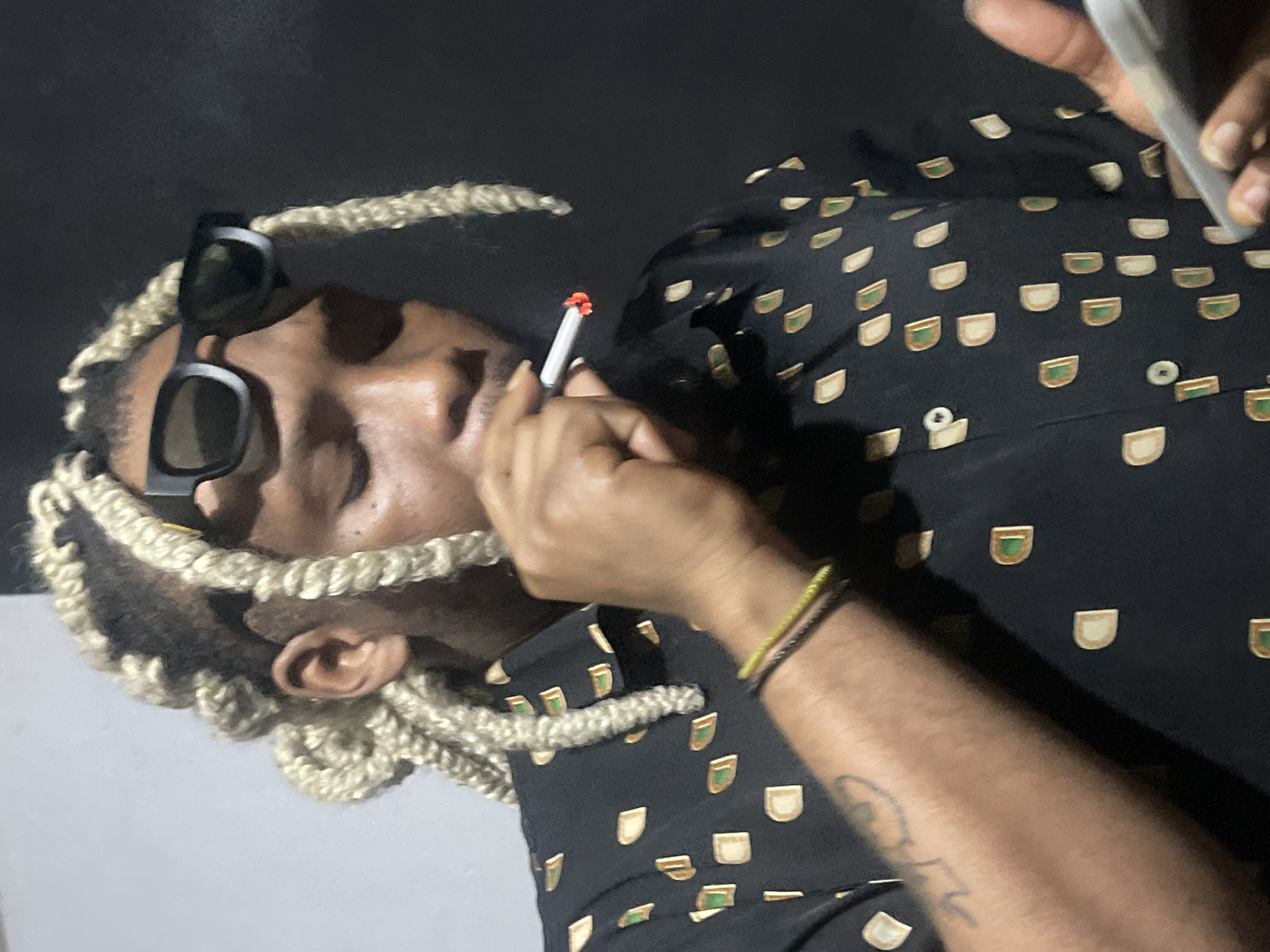A person with blonde braided hair and black sunglasses is smoking a cigarette. They are wearing a black shirt with a repeating pattern of tan and green shapes. A tattoo is visible on their left arm. The background is dark, suggesting an indoor setting or nighttime. The image shows no specific indicators of being in Jos, Nigeria.