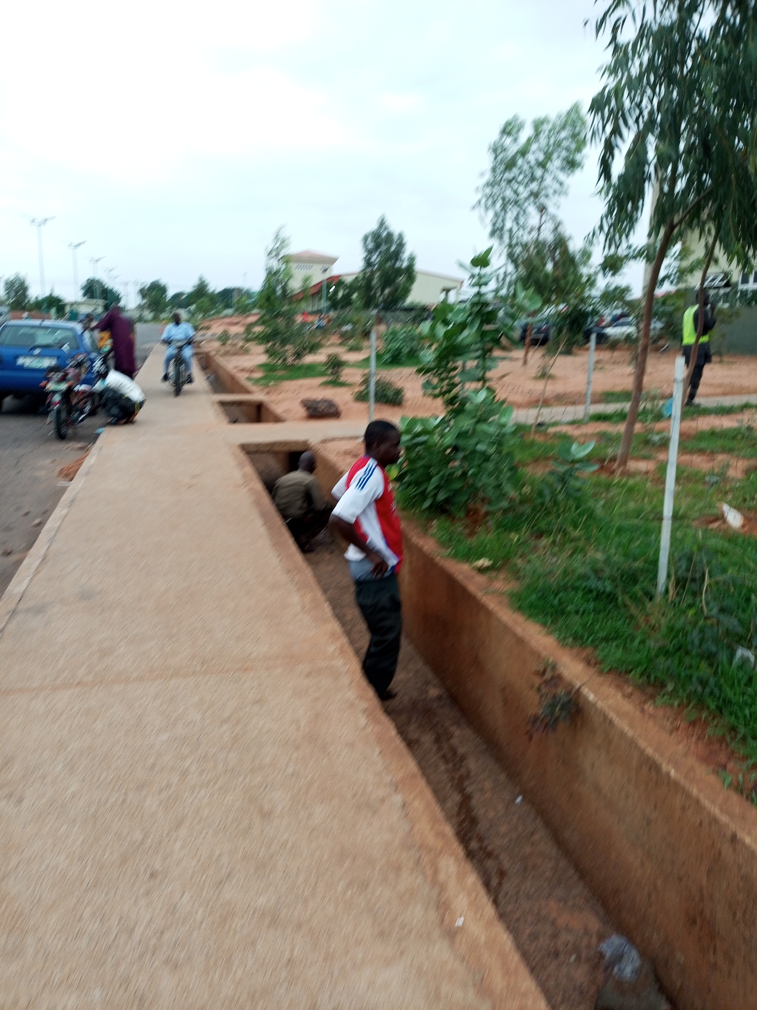 The image captures a street scene in Gombe, Nigeria, with a focus on a concrete drainage channel. In the foreground, a man in a red and white shirt and black pants stands near the edge of the drainage ditch. Further down the ditch, another man wearing a khaki shirt and dark pants is partially visible. On the sidewalk, a blue car is parked, and a motorcycle is also present. A person in a dark maroon robe stands near the parked car, and another individual is seen on a bicycle in the background. The environment appears to be an urban setting with buildings and trees visible in the distance. The sky is overcast, suggesting a cloudy day. The ground is a mix of dirt and sparse vegetation.