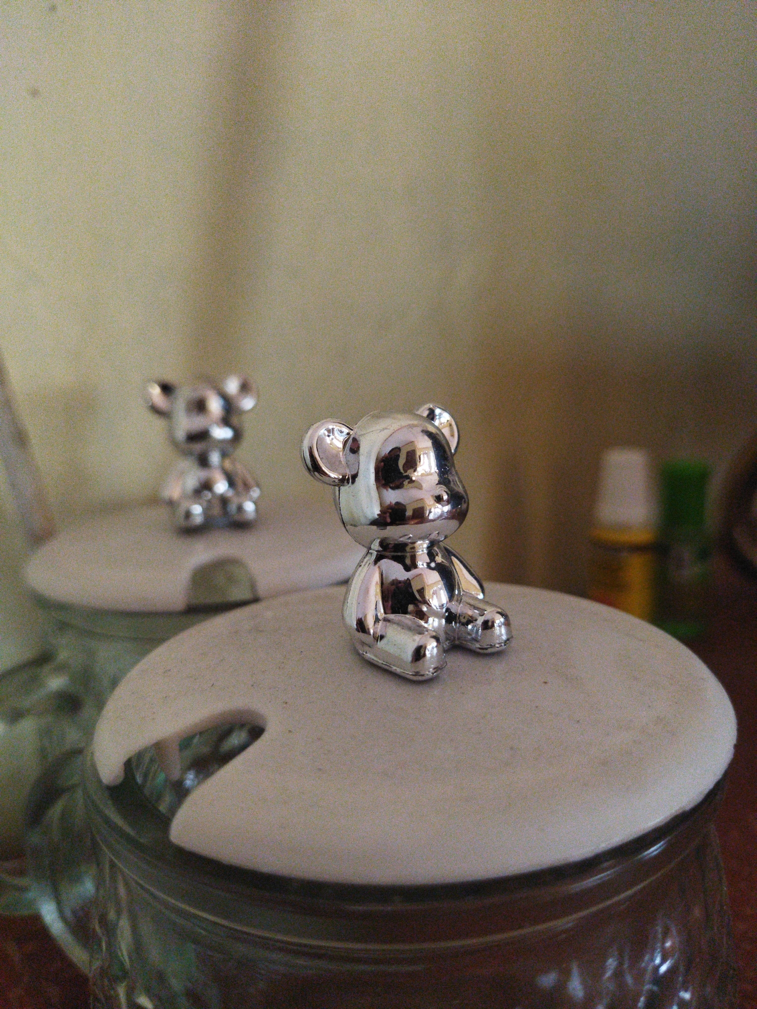 The image displays two small, shiny, silver bear figurines positioned on top of two jars. The bear in the foreground is larger and in sharper focus, while the second bear, further in the background, is smaller and out of focus. Both bears are sitting upright. The jars have light-colored lids with cutouts. The background is a muted, textured surface, possibly a wall, with some out-of-focus bottles visible on the right. The overall lighting is soft. There is no discernible information within the image that would indicate a location in the city of Suci, Indonesia, or any other specific geographical area.