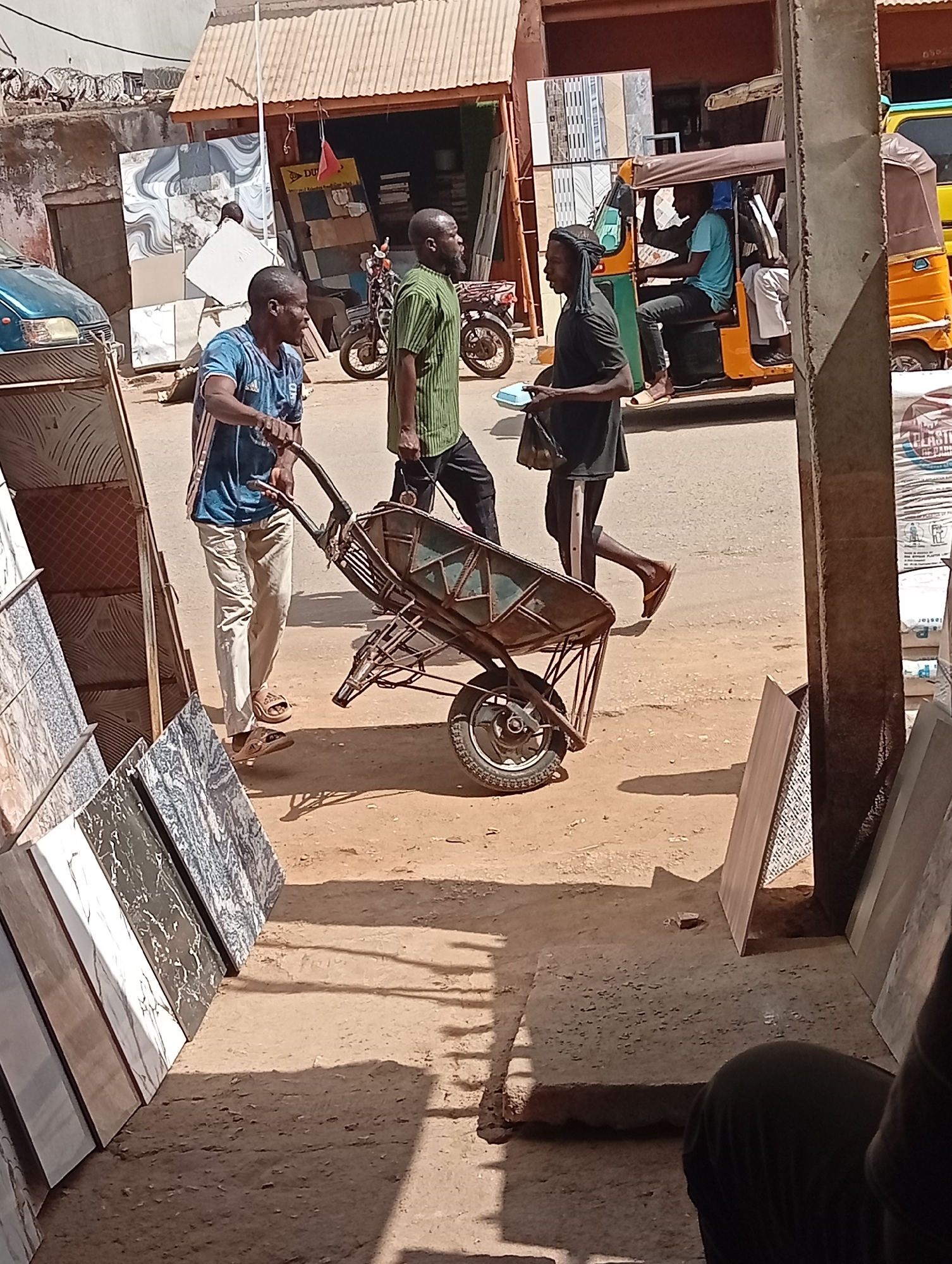 This media file captures a bustling street scene, likely in a market or commercial area, in Maiburiji, Nigeria, under bright daylight. The ground is dusty earth, indicating dry weather.
In the foreground, a man in a blue and white striped t-shirt and light-colored trousers pushes an empty metal wheelbarrow with a single wheel. He wears brown sandals. Beside him, another man in a green and black striped shirt and dark trousers walks with what appears to be a bag in his left hand, facing away from the viewer. A third man, dressed in a black t-shirt and dark trousers with a white stripe, walks in the opposite direction, holding an item in his hand, possibly a small tray or book. He wears light-colored sandals. His head is wrapped in a dark fabric.
The street is lined with commercial establishments. To the left, numerous ceramic tiles of various patterns and sizes are displayed upright, leaning against a rack and structures, suggesting a tile shop. A blue car is partially visible behind these displays. Further in the background, another shop displays more tiles. A yellow sign with "DU" (possibly part of "DUNLOP") is visible above one of the shop entrances.
On the right side of the street, an orange and green tricycle (known as a "keke" in Nigeria) is parked or moving slowly. Two individuals are visible inside: one at the wheel, wearing a light blue t-shirt, and another passenger in white attire. Behind the tricycle, bags of material, some labeled "PLASTER," are stacked. A motorcycle is also visible between the shops and the tricycle. Strong shadows indicate a clear, sunny day. The overall scene depicts typical daily life and commerce in a West African town.