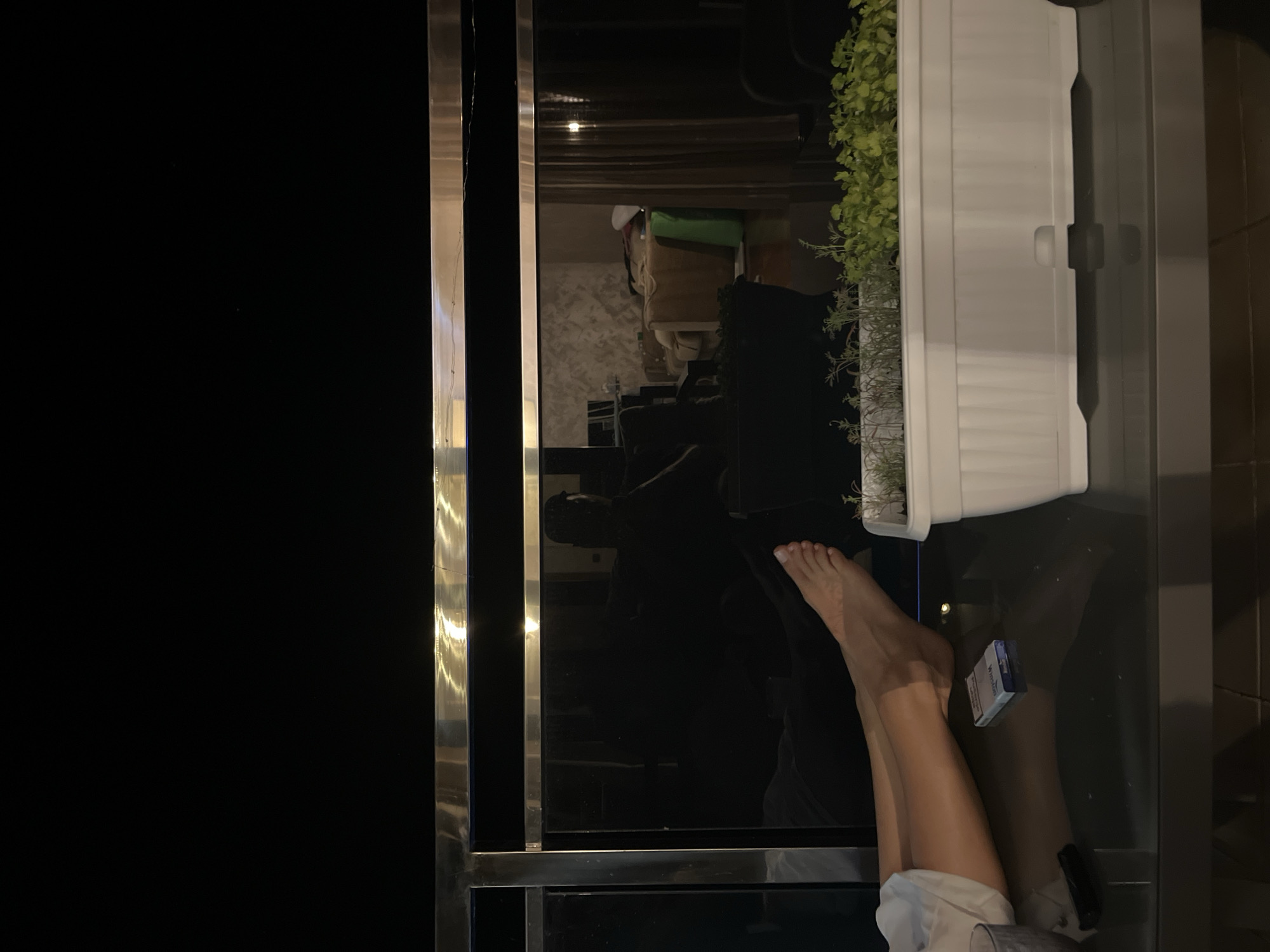 The image depicts a close-up of a person's legs and feet resting on a glass table, with a pack of cigarettes in front of them. In the background, a large black screen, possibly a television or window, reflects a room interior. A white planter filled with greenery sits on the table. The scene appears to be set on a balcony or patio in Budva, Montenegro, as suggested by the presence of a balcony railing in the foreground and the overall ambiance. The lighting is dim, with some reflections from artificial sources. The overall impression is one of a relaxed evening setting.
