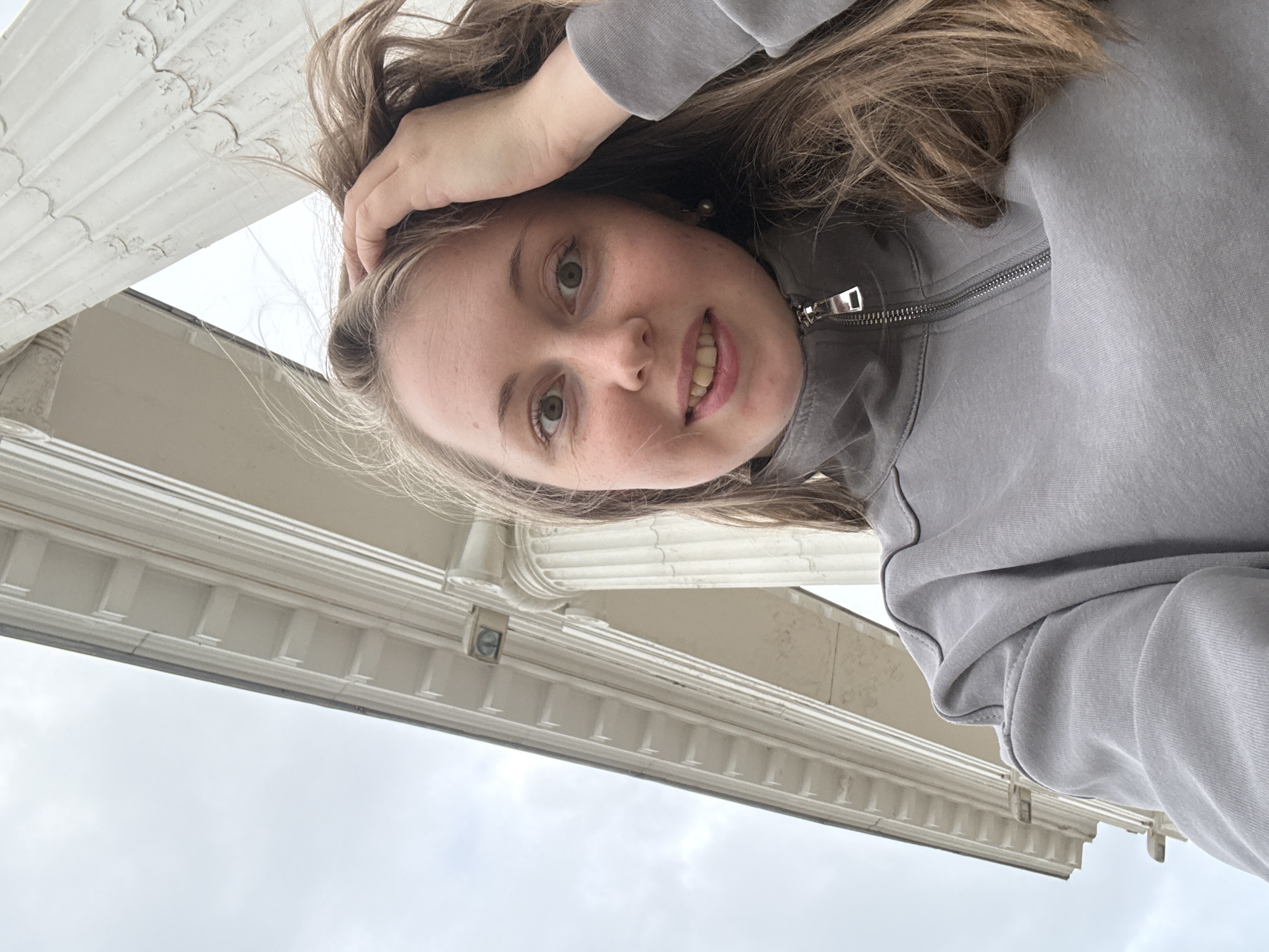 A young adult female takes a low-angle selfie in front of a classical or neoclassical building under an overcast sky. She has fair skin, light brown hair, and light-colored eyes, looking directly at the camera with a slight smile. Her left hand is raised, with fingers in her hair near her head. She wears a grey, long-sleeved quarter-zip sweatshirt with a metallic zipper. A pearl-like earring is visible in her right ear. The background features large, white architectural elements, including fluted columns and an entablature with distinctive rectangular blocks (possibly dentils or modillions). The building material appears to be painted stone or concrete, showing some signs of wear and minor peeling on the columns. A dark, rectangular object, possibly a light fixture or sensor, is embedded in the entablature above the column on the left.