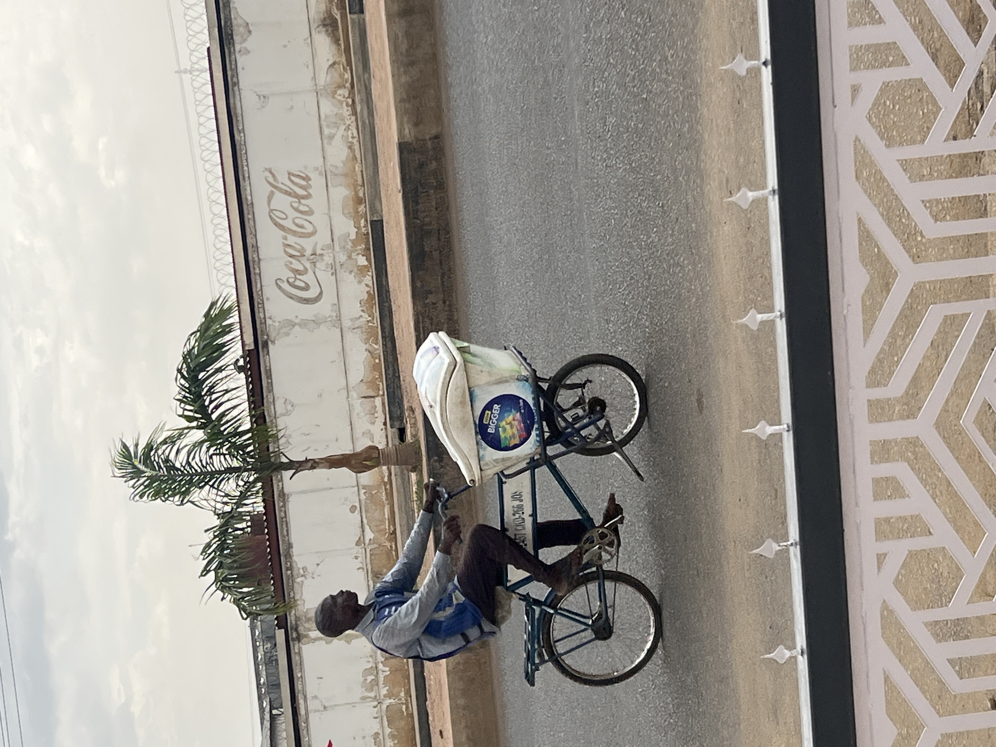 A man rides a three-wheeled bicycle with a cooler on the front along a road in Barakin Lerawi, Nigeria. A Coca-Cola sign is on a building behind a palm tree. A decorative fence is in the foreground.