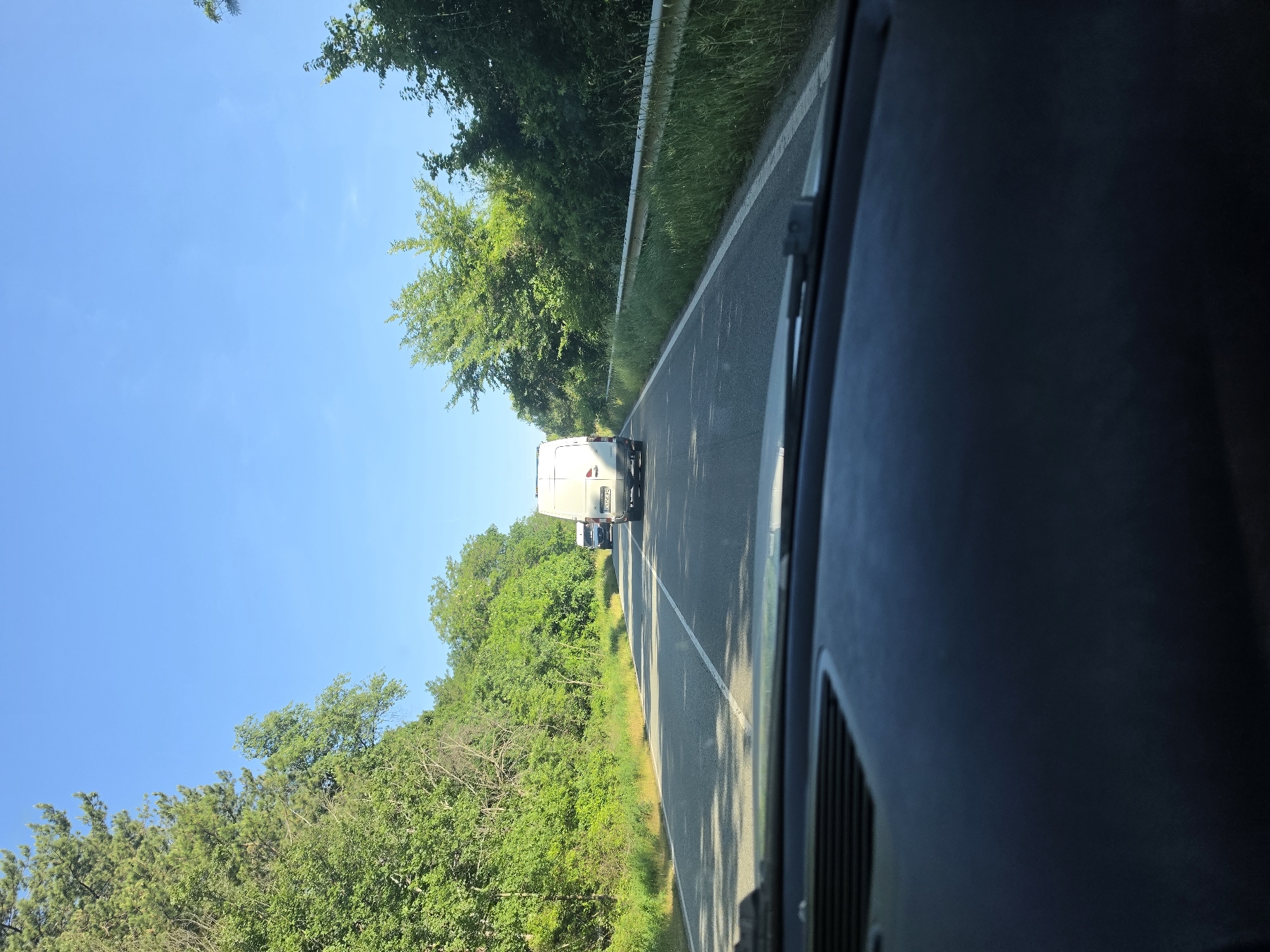 The image is a forward-facing view from inside a vehicle on a two-lane road bordered by lush green trees and vegetation. The sky is clear and blue. In the distance, two white vans are traveling in the same direction. The closest van is a large box van, and behind it is a smaller van. The road has a white line marking the edge and a dashed white line dividing the lanes. The foreground shows the dark dashboard of the vehicle from which the photo was taken. The scene appears to be a rural or suburban setting. The location is identified as Blatino, Bulgaria.