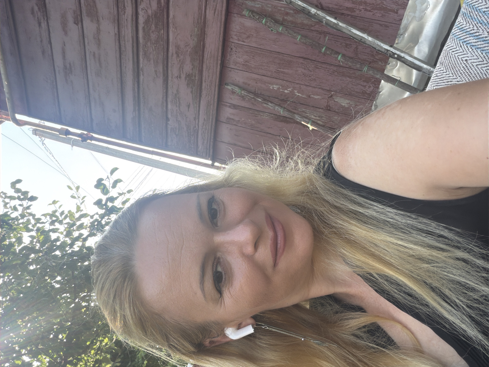 A woman with blonde hair, wearing a black top and white earbuds, poses for a selfie outdoors. She is positioned in the foreground, with a weathered wooden wall and some vegetation in the background. The lighting suggests it is daytime, possibly sunny, with some lens flare visible. The woman has makeup on, and her expression is a gentle smile. The wooden wall is a deep red-brown color and appears to be part of an outdoor structure, possibly a shed or fence. There are some vertical wooden stakes leaning against the wall, bound with green twine. The environment seems rustic and somewhat overgrown.