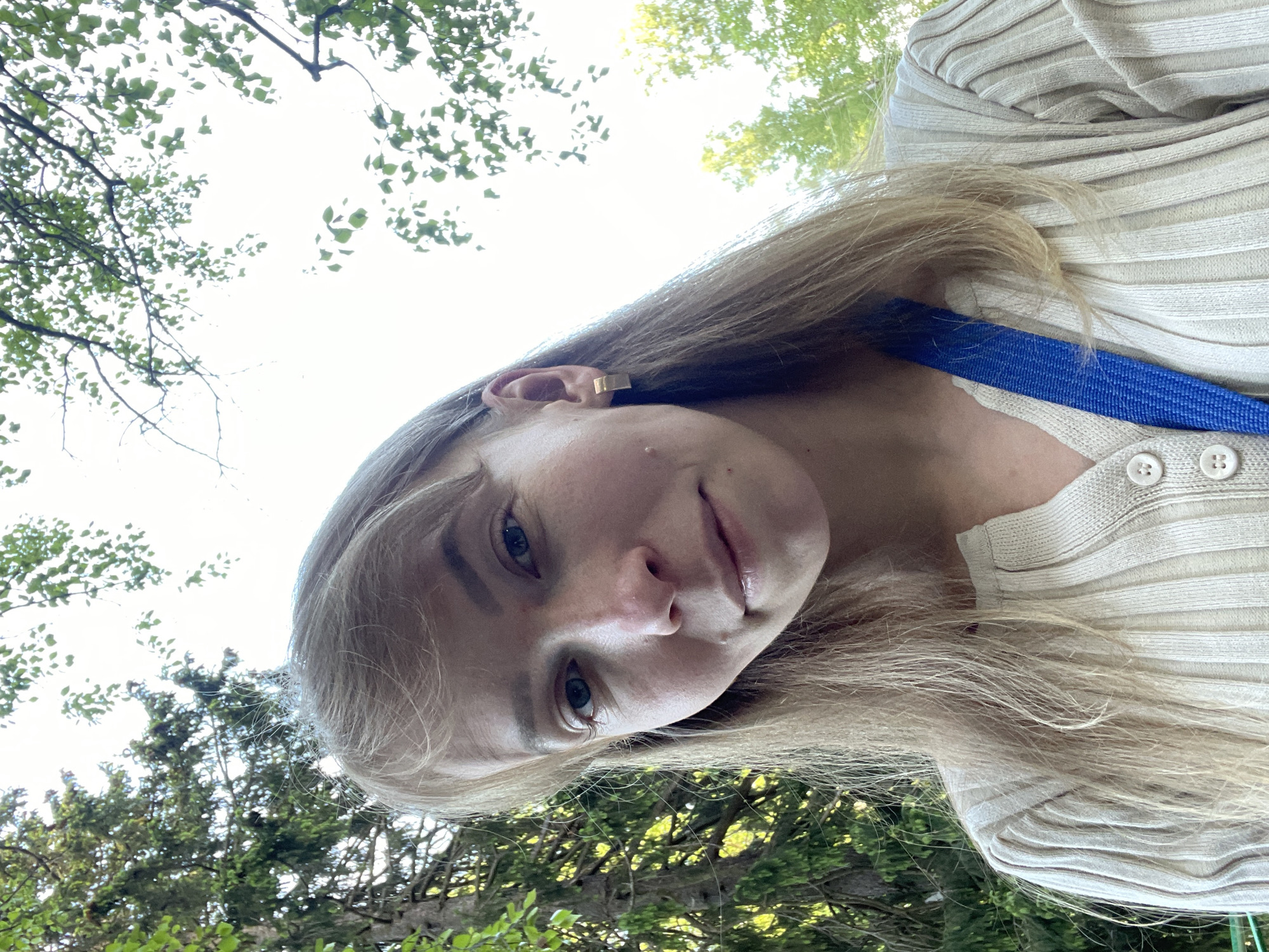 A young, blonde woman is photographed from a low angle, looking down at the camera. She has fair skin and is wearing a light-colored, ribbed cardigan with white buttons. A blue strap is visible across her shoulder. She is wearing a gold-colored earring on her left ear. The background shows green trees and sky, suggesting she is outdoors. The location in the city of Perm, Russia is not ascertainable from visual information.