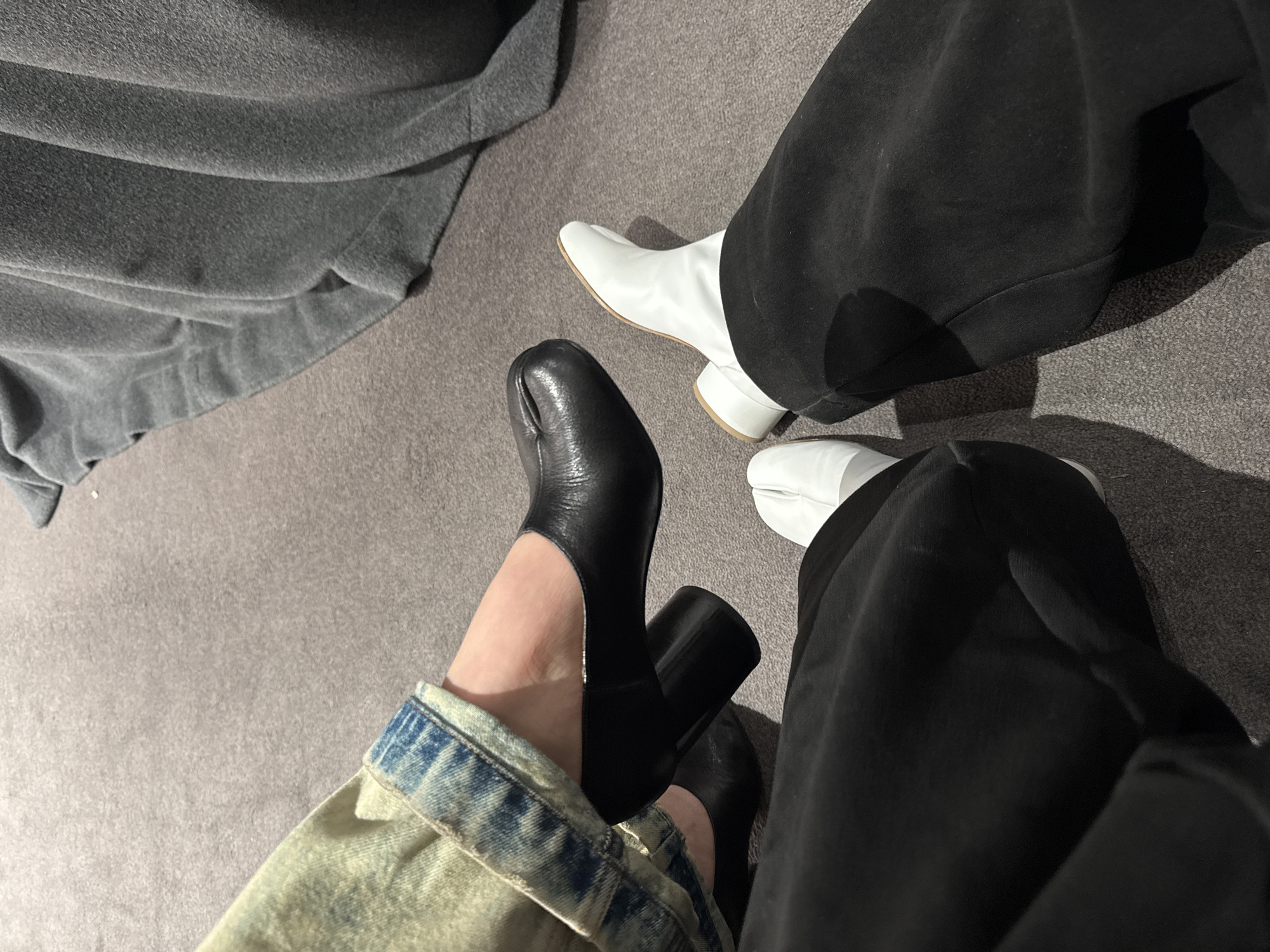 An overhead shot shows two pairs of feet, one wearing black Tabi heels and the other wearing white Tabi boots. The foot wearing the black heels is clad in a distressed denim jacket cuff. The other foot is wearing baggy black pants. A gray fabric is draped in the upper left corner of the frame. The floor is gray carpet. There is no indication of the location being Paris, France.