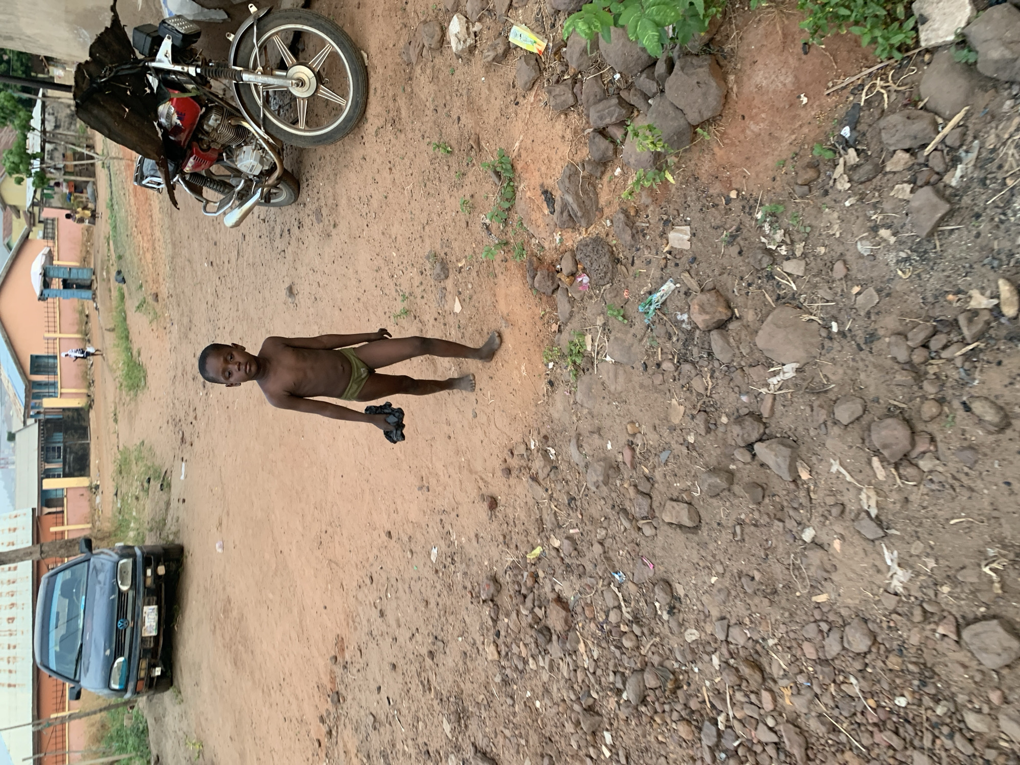 The image shows an outdoor scene in Lokoja, Nigeria. A young boy stands shirtless in green underwear in the middle of a dirt road. He holds a dark object in his left hand. To the left of the boy, a blue car is parked. Further up, a motorcycle is parked next to the road. The road surface varies from compacted dirt to rocky patches with sparse vegetation. Litter is scattered across the ground. Buildings are visible in the background.