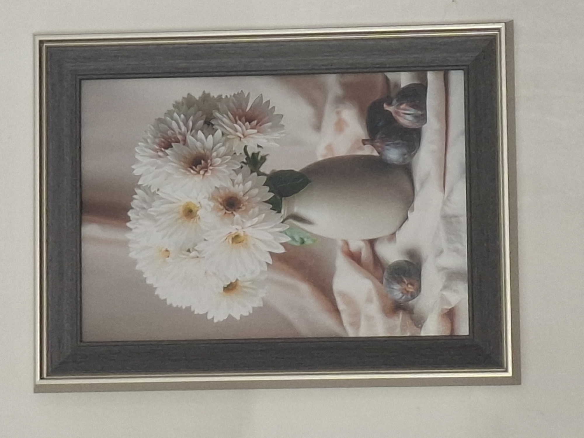 The image is a photograph of a framed still life painting. The painting depicts a bouquet of white dahlias with pinkish centers in a beige vase. The vase sits on a draped pinkish fabric and a white cloth. Three figs are placed in the foreground on the white cloth. The painting is framed by a dark gray mat and a metallic silver frame. The photograph is taken at a slight angle, showing the painting on a light-colored wall. The location in Almaty, Kazakhstan, is not discernible from the image itself.