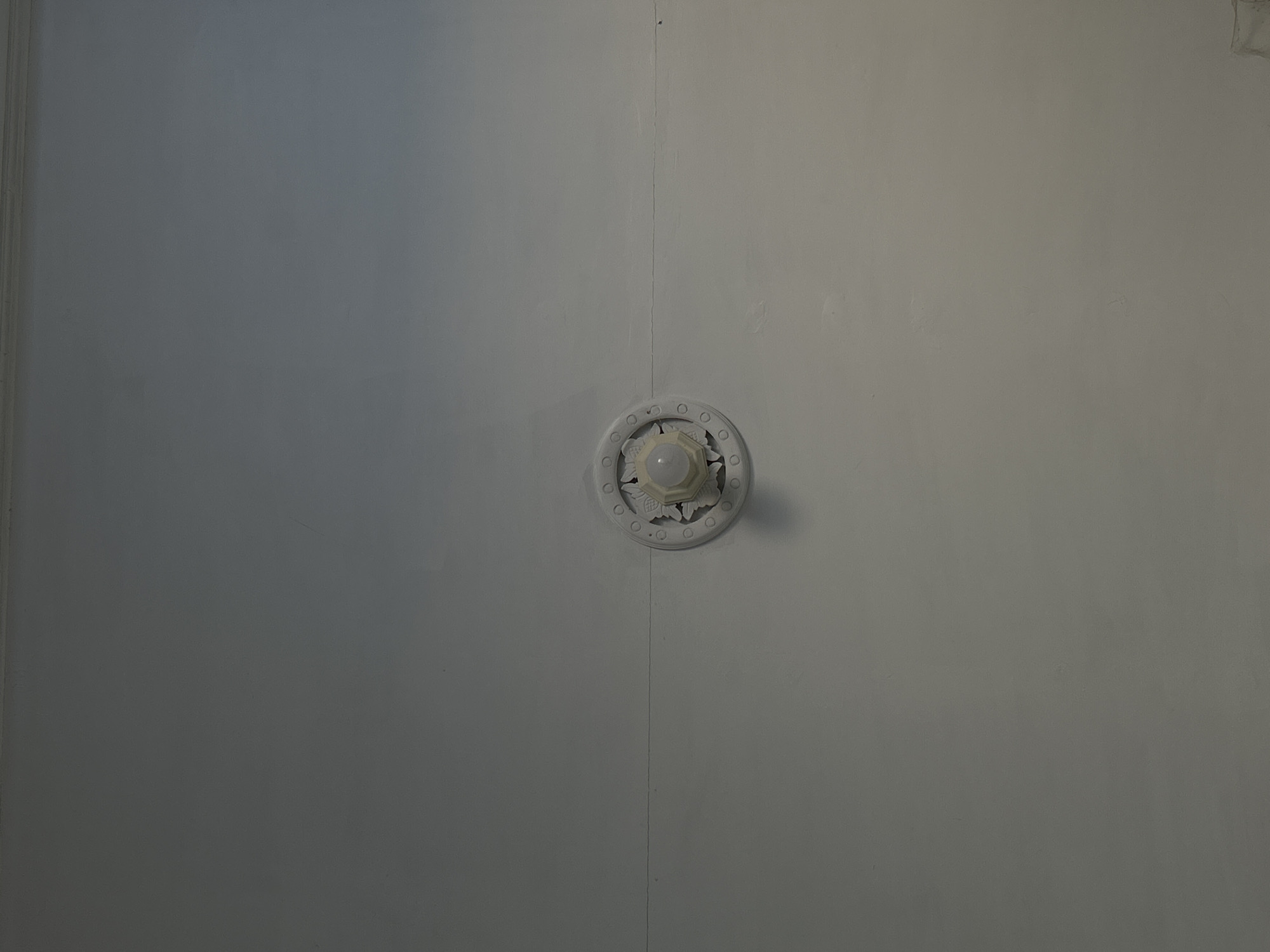 The image depicts a close-up of a white, seemingly painted metal or plaster surface. A circular light fixture is centrally located, featuring a decorative floral pattern around its edge. The bulb is visible within the fixture, appearing somewhat dim or covered. A vertical seam or joint runs through the center of the image, suggesting that the surface is composed of multiple panels. There is no information to indicate the location is in Lhokseumawe, Indonesia.