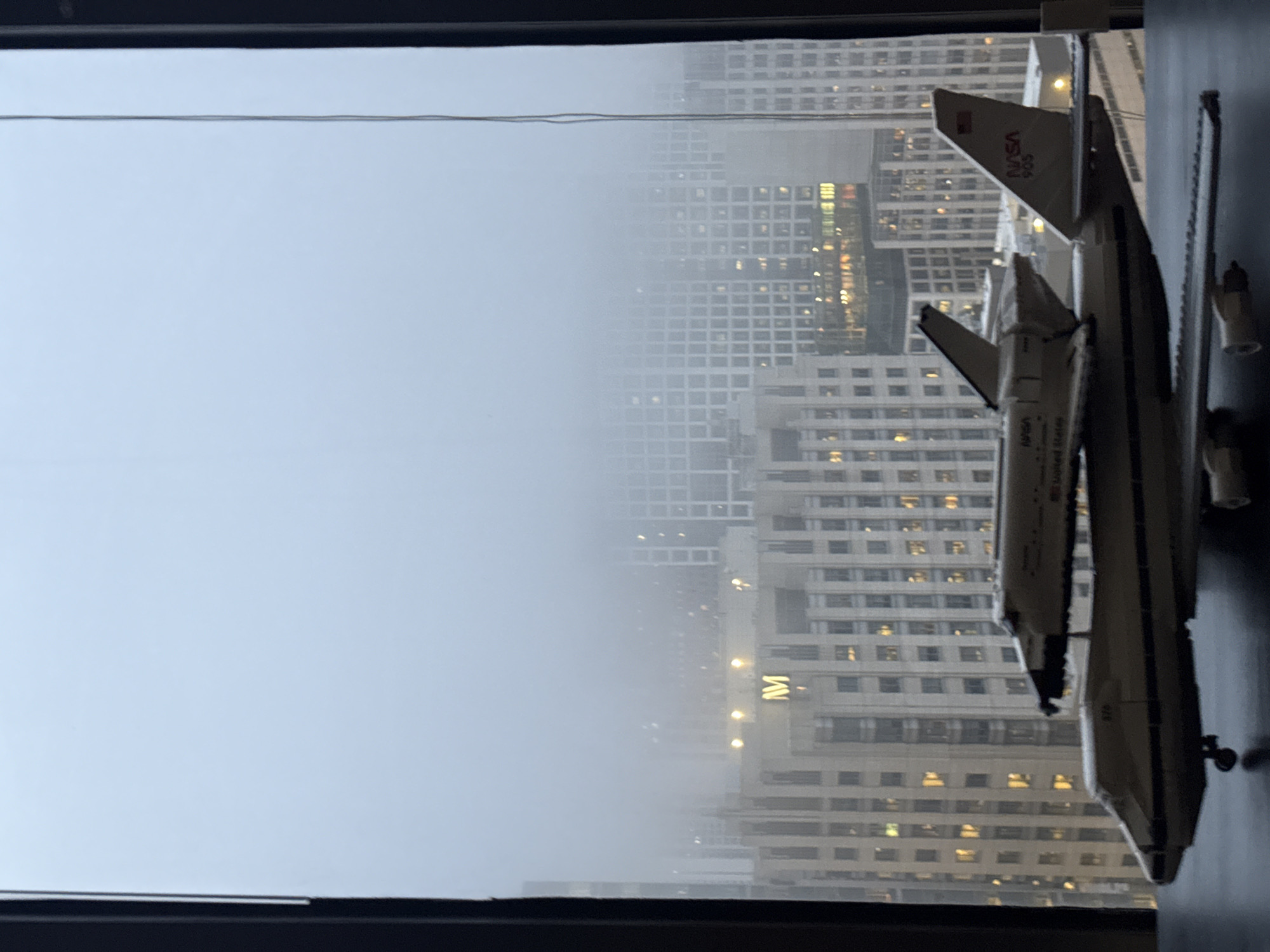 The image shows a model of a space shuttle mounted on top of a model airplane. The models are in the foreground, placed on a dark surface. In the background, a cityscape is visible through a window. The city is shrouded in fog, obscuring many of the buildings. A large building in the mid-ground features illuminated lights and a prominent "M" logo, suggestive of the Chicago Merchandise Mart. The overall atmosphere is hazy and muted due to the fog. No people are visible in the image.