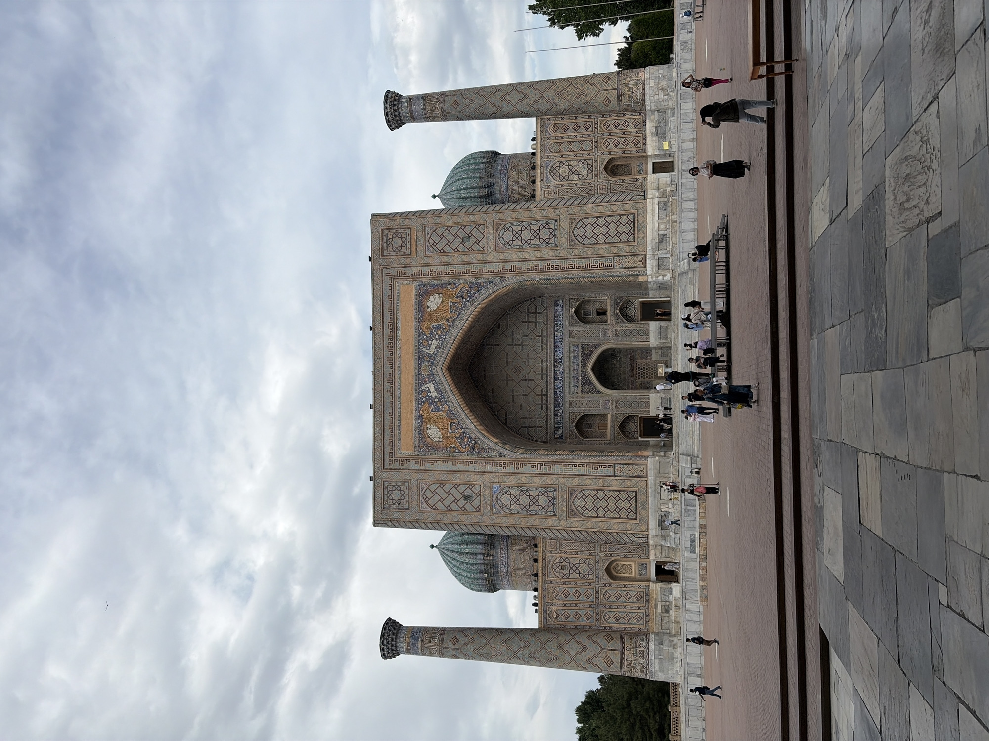 A grand architectural complex, likely the Sher-Dor Madrasah, dominates the scene under an overcast sky in Samarkand, Uzbekistan. The ornate building features a towering central pishtaq (archway), flanked by two tall, intricately tiled minarets at its corners. The facade is a breathtaking display of Islamic artistry, adorned with elaborate geometric patterns, floral motifs, and detailed calligraphic script rendered in various shades of blue, turquoise, brown, and white tiles.
A distinctive feature is the vibrant depiction on the central arch, showing a lion-like creature with a human face surrounded by a sun, a unique characteristic of this madrasah. Two domes are visible, one prominently above the central arch and another partially seen to the right, complementing the majestic structure.
In the foreground, a wide, paved plaza extends before the building. The ground is covered in large grey stone tiles on the right, transitioning to a reddish-brown tiled area closer to the monument. Several people are scattered across this plaza, walking or standing, suggesting a public space for visitors and tourists. Green foliage from trees is visible at the left and right edges of the frame.
The weather appears cloudy, indicating a mild daytime setting. Visible text on the building's facade, identified through OCR, includes "الله", "الحمد لله", and "لا إله إلا الله والله أكبر", indicating religious inscriptions.