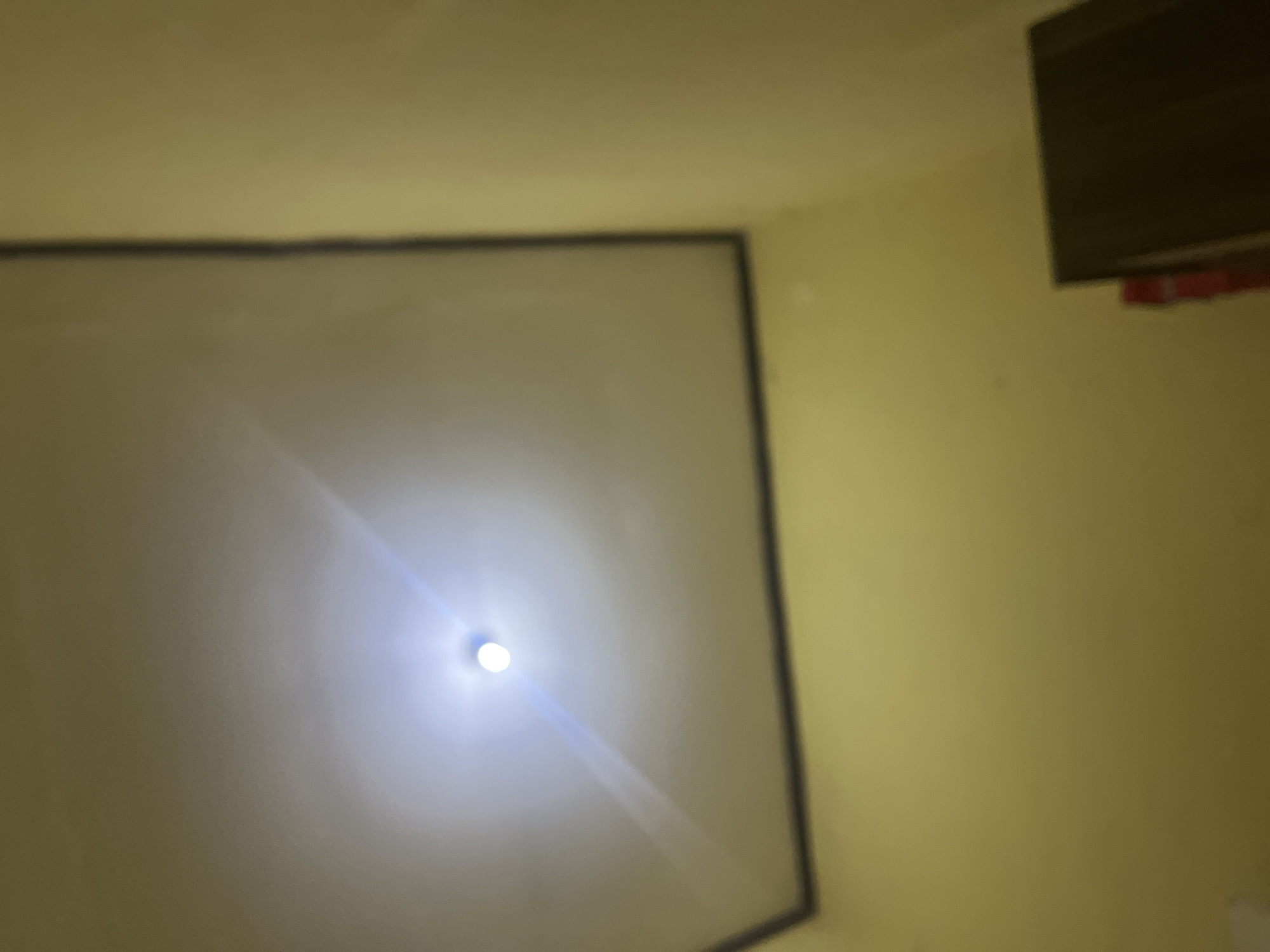 The image displays an interior view of a room, likely a residential or office space. A ceiling light is prominently visible in the center of the image, emitting a bright, diffused light that creates a halo effect. The ceiling appears to be a flat, recessed panel, bordered by a darker trim. The walls are painted a light yellow or cream color. In the lower right corner of the image, a dark rectangular object, possibly a piece of furniture like a cabinet or shelf, is partially visible. The overall impression is one of a simple, functional room, dimly lit except for the direct illumination from the ceiling fixture. There are no people or overt actions depicted in the image. The location of Cirahayu, Indonesia, is not directly ascertainable from the visual information presented in the photograph.