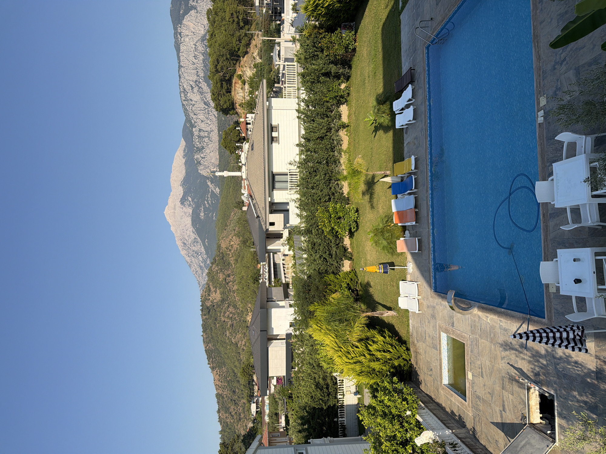 This is a high-angle exterior shot of a residential property with a view of mountains in the distance. A large, rectangular swimming pool with blue water occupies the foreground, bordered by a tiled patio. Several lounge chairs are arranged on the patio, with some in the sun and some in the shade. A patio table with chairs is also visible. Beyond the pool and patio, there is a well-maintained lawn surrounded by lush green hedges and trees. Several modern houses with dark roofs are visible behind the hedges, and further in the distance, a mosque with a tall, slender minaret stands out against the backdrop of rolling, tree-covered hills and a prominent, rocky mountain peak. The sky is clear and bright blue, indicating fair weather. The overall scene suggests a tranquil, upscale residential area in a mountainous region.