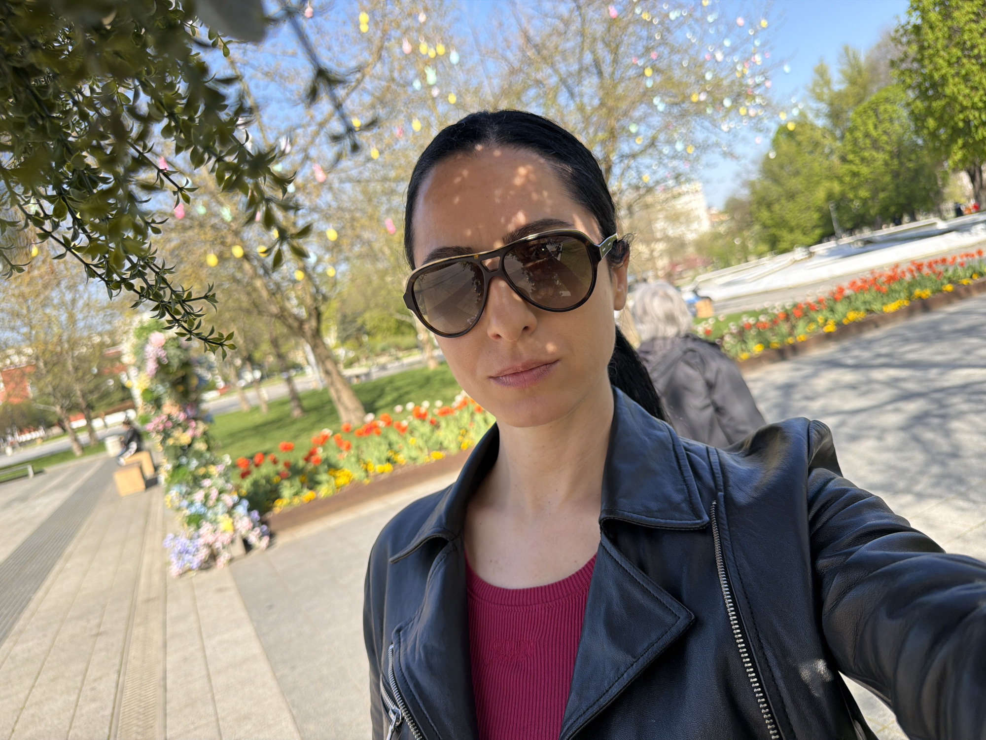 A woman takes a close-up selfie outdoors on a sunny day in Pleven, Bulgaria. She is positioned slightly to the right, looking directly into the camera with a neutral expression. She wears large, dark aviator-style sunglasses with a gold-colored bridge, a black leather jacket, and a dark pink or red ribbed top with a subtle, possibly embroidered, logo near the neckline. Her dark hair is pulled back, and dappled shadows from overhead foliage fall across her face and forehead.
The background reveals a vibrant spring setting. Numerous deciduous trees line a paved pedestrian area, their bare branches adorned with colorful, egg-shaped decorations in yellow, pink, and light blue, suggesting an Easter or spring festival. Lush flower beds with bright red and yellow tulips and other colorful blooms are visible behind her, bordering a raised, white structure, possibly part of a fountain or decorative landscape element. Another person is partially visible in the mid-ground, facing away from the camera. Overhanging green foliage partially frames the top left of the image. The sky is clear and blue, indicating bright daylight, likely mid-morning or afternoon. The scene depicts a lively and decorated urban park or square.