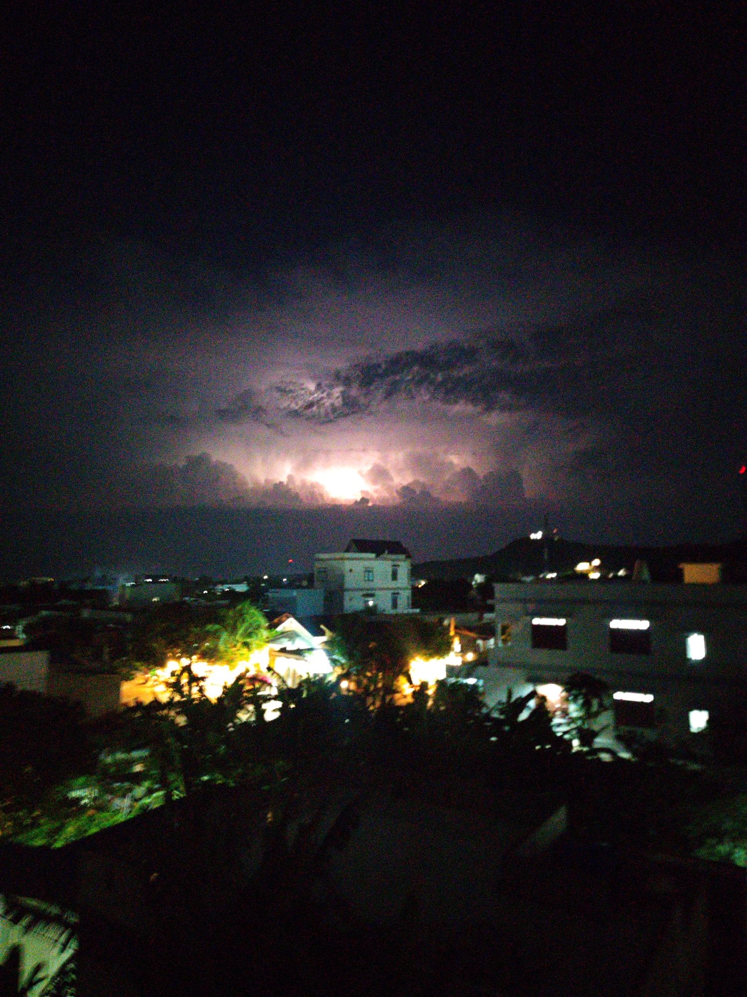Captured at night in Phú Quý, Vietnam, the image reveals a city skyline under a turbulent sky. In the distance, a mountain rises slightly above the horizon. A burst of light, likely lightning, illuminates the cloud cover. Buildings dot the cityscape, some with illuminated windows, creating bright spots amidst the darkness. Trees are visible in the foreground.