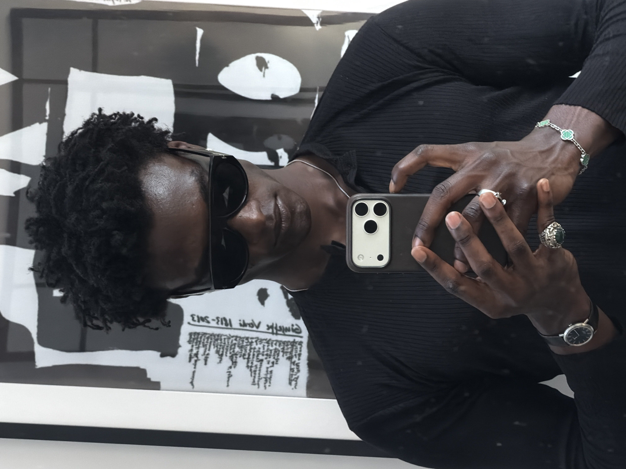 A person with dark skin, curly black hair, and wearing black sunglasses and a black ribbed shirt is taking a selfie with a cell phone in front of artwork. The phone has three camera lenses. The person is wearing a silver necklace, a bracelet with green quatrefoil charms, a watch with a black band, and multiple rings. The artwork behind them has abstract shapes in black and white, and some text at the bottom. The person appears to be in a gallery or museum setting. The location is likely in London, United Kingdom.