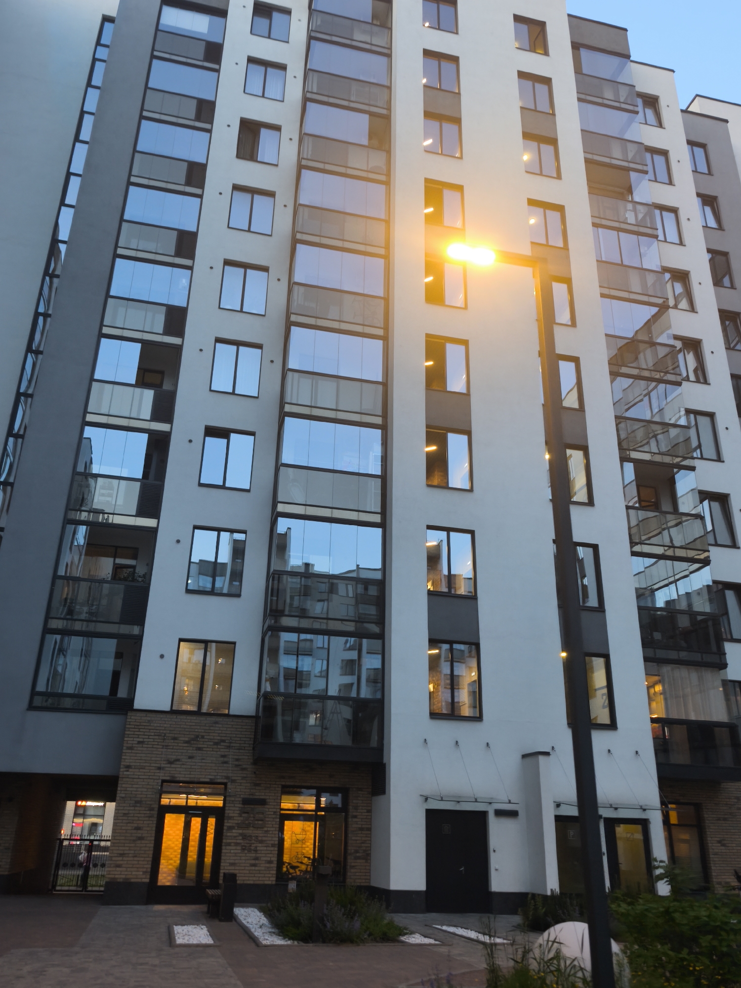 This image depicts the exterior of a modern residential building, likely an apartment complex, in Saint Petersburg, Russia. The building is tall and features a predominantly white facade with dark gray accents. Numerous rectangular windows are visible, many of them reflecting the sky and surrounding environment, which appears to be twilight or dusk. Some windows are lit from within, indicating occupancy. The building has a series of balconies with glass railings.
The lower section of the building includes an entrance area with brick detailing around doorways. One entrance features a glass door with a warm yellow light emanating from within, illuminating the brickwork. A black lamppost with a brightly lit lamp stands in front of the building, casting a strong yellow-orange glow onto the facade.
The ground in front of the building is paved with interlocking bricks. There is a small landscaped area with some greenery and white decorative stones. A black metal fence is visible to the left of the entrance area, partially obscuring a glimpse of what might be a street or pathway beyond. The overall impression is of a contemporary urban residential structure.