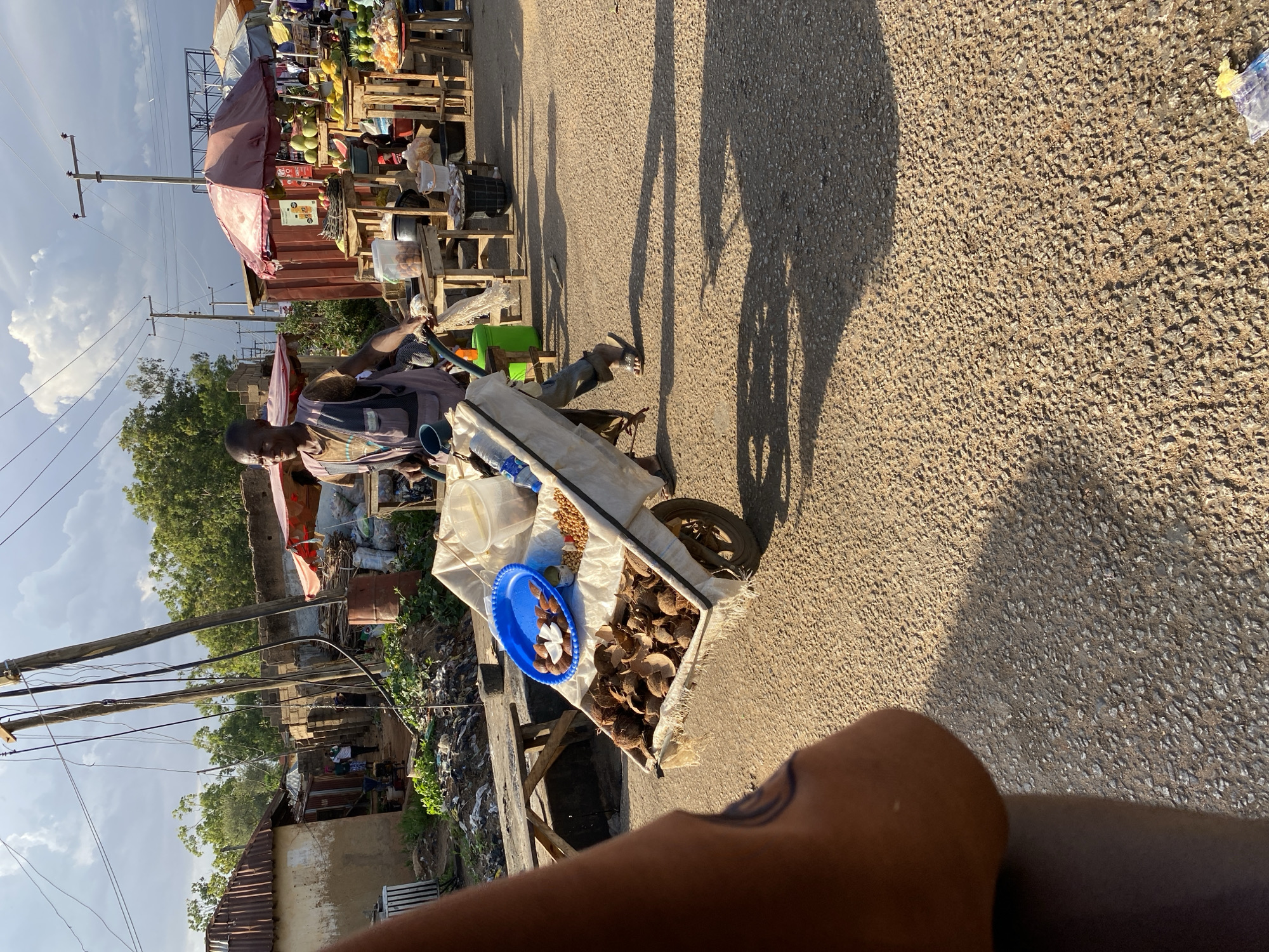 The image captures a street scene in Bukuru, Nigeria. An elderly man pulls a cart containing coconuts and peanuts along a paved road. The road appears to have other vendors and market stalls set up along it, selling goods. Shadows of people and objects are cast onto the road, suggesting a sunny day. A residential area or slum is present on the left side of the image. Power lines are visible against the sky. The image is taken from a first-person perspective, with part of the photographer's arm visible in the foreground.