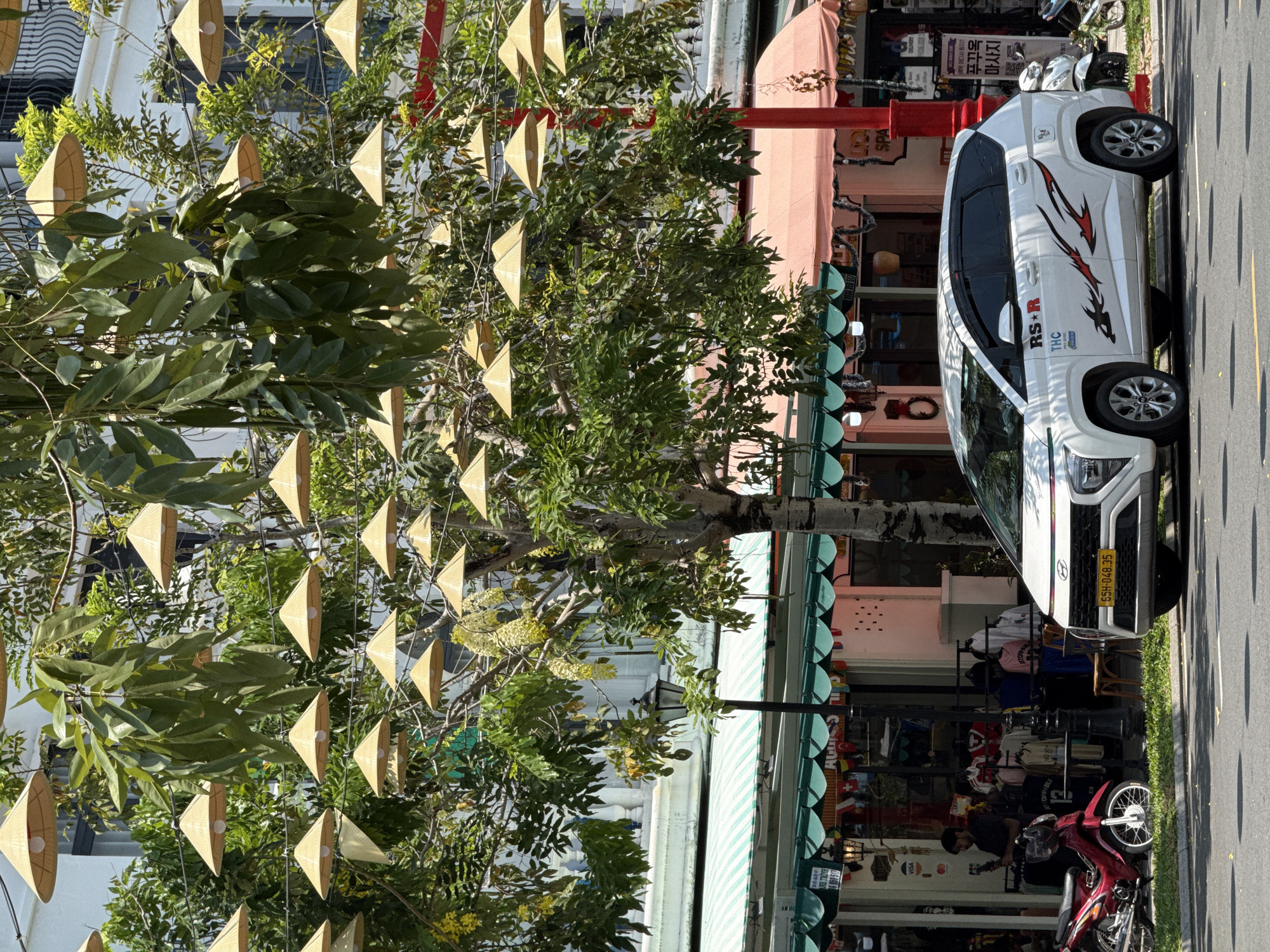 The image depicts a vibrant, sunny street scene, likely in a commercial area of Phú Quốc, Vietnam. The time of day appears to be midday or early afternoon due to the bright light and distinct shadows.
In the left foreground, several trees are heavily adorned with numerous small, conical-shaped decorations, resembling traditional Vietnamese nón lá hats, hanging from their branches. One of the trees also features clusters of light yellow flowers.
To the right, a white car, possibly an SUV, is parked along the street. It features custom decals on its side, including red and black stylized graphics, the text "RS★R", and "THC". Its yellow Vietnamese license plate reads "65H-048.35". Further along the curb, a red moped is parked, and a person is partially visible near it, seemingly interacting with the shopfront. Another white moped is visible further down the street.
Behind the vehicles and trees are several shopfronts with awnings. One awning is a prominent pink/salmon color, while another features green and white stripes. Merchandise is displayed in the shop windows. A red pillar, possibly a decorative element or mailbox, stands between the shops.
Visible text in the scene includes: "ANH TRINH", "VISA", "TOPPABLE", "STOPPA", "DELIVERY", and "SPA". There is also Korean text on a sign, which translates to "예약 어디서 하지?" (Where to make a reservation?), "푸꾸옥" (Phú Quốc), and "마사지" (Massage), reinforcing the location and available services. The "nón lá" decorations, along with the Vietnamese license plate and mixed language signage, strongly indicate a Southeast Asian setting, specifically Vietnam.