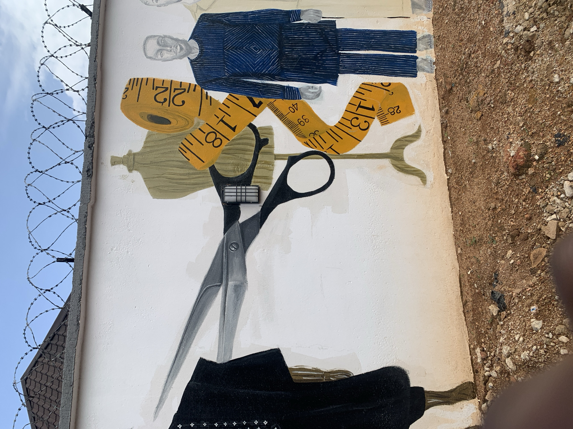 This is a mural on an outdoor wall in Gura Top, Nigeria. The mural depicts elements related to tailoring and fashion. In the foreground, there is a black garment on a mannequin to the left, and a pair of large scissors cutting across a yellow measuring tape. To the right of the measuring tape, there are two figures dressed in traditional Nigerian attire. The figure on the left is wearing a blue patterned outfit, and a portion of another figure in a light-colored outfit is visible to the far right. Behind the mural, a white wall extends upwards to a concrete ledge topped with coiled barbed wire under a clear blue sky with some clouds. The ground in front of the wall is covered in reddish-brown dirt with scattered rocks and debris.
