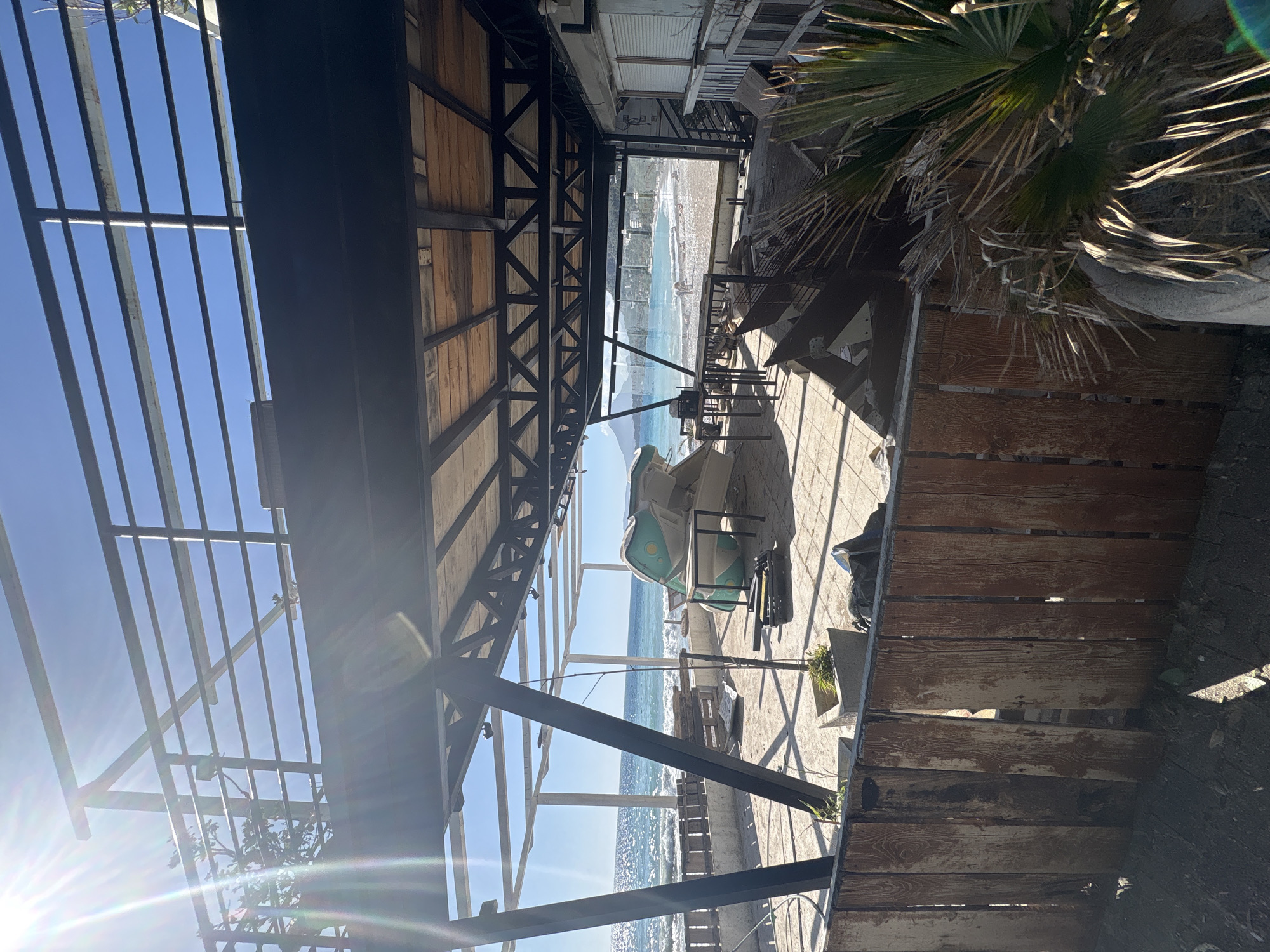 The image captures a view from under a partially constructed or dilapidated metal and wood structure, likely a canopy or pergola, looking out towards the beach and the sea in Bar, Montenegro. The perspective is slightly skewed, as if the camera is tilted.
The foreground is dominated by the structure, with its metal beams and wooden slats creating a grid-like pattern. Some of the wood appears aged and worn. To the right, there's a section of a weathered wooden fence or barrier, and a palm tree.
Beyond the structure, the scene opens up to a paved area with some outdoor furniture, including chairs with light green cushions. Further in the distance is the beach, with waves lapping at the shore. The sea extends towards the horizon, where the sky is visible, with some hazy or bright light conditions. It appears to be a sunny day. The image conveys a sense of being in a transitional space between the built environment and the natural landscape of a coastal area.