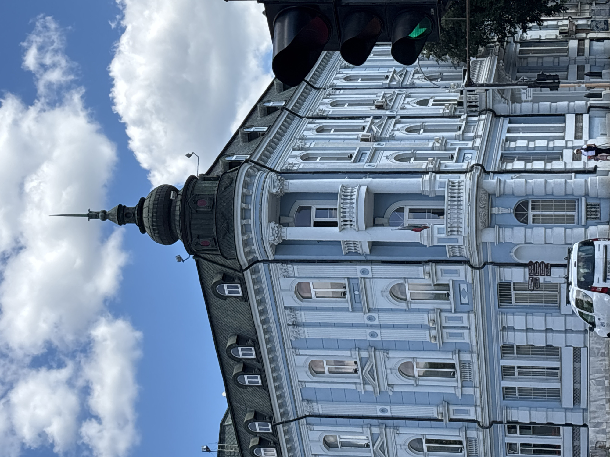 The image shows the exterior of a light blue building with ornate architectural details, located in Varna, Bulgaria. The building features multiple windows of various shapes and sizes, classical columns, and a domed turret topped with a spire. A dark green roof with dormer windows is visible. In the foreground, a traffic light with a green signal is positioned to the right of the building. A white car is parked on the street below the building. Two individuals, one adult and one child, are walking on the sidewalk. The sky is blue with scattered white clouds.