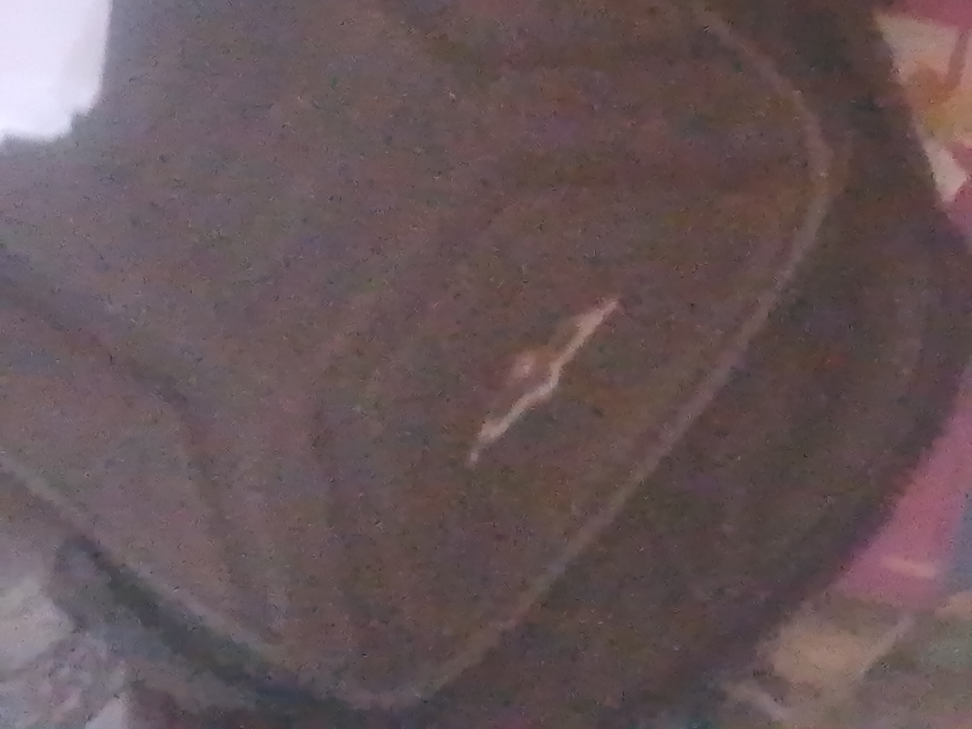 The image is a close-up, blurry shot of a dark, textured surface. A faint, light-colored, V-shaped mark is visible in the center. The texture of the surface appears to be fabric or rough material. The background is out of focus and contains indistinct shapes and colors, possibly other objects or a wall. There is no identifiable location in Jauro Yinu, Nigeria, or any discernible people, actions, or interactions.