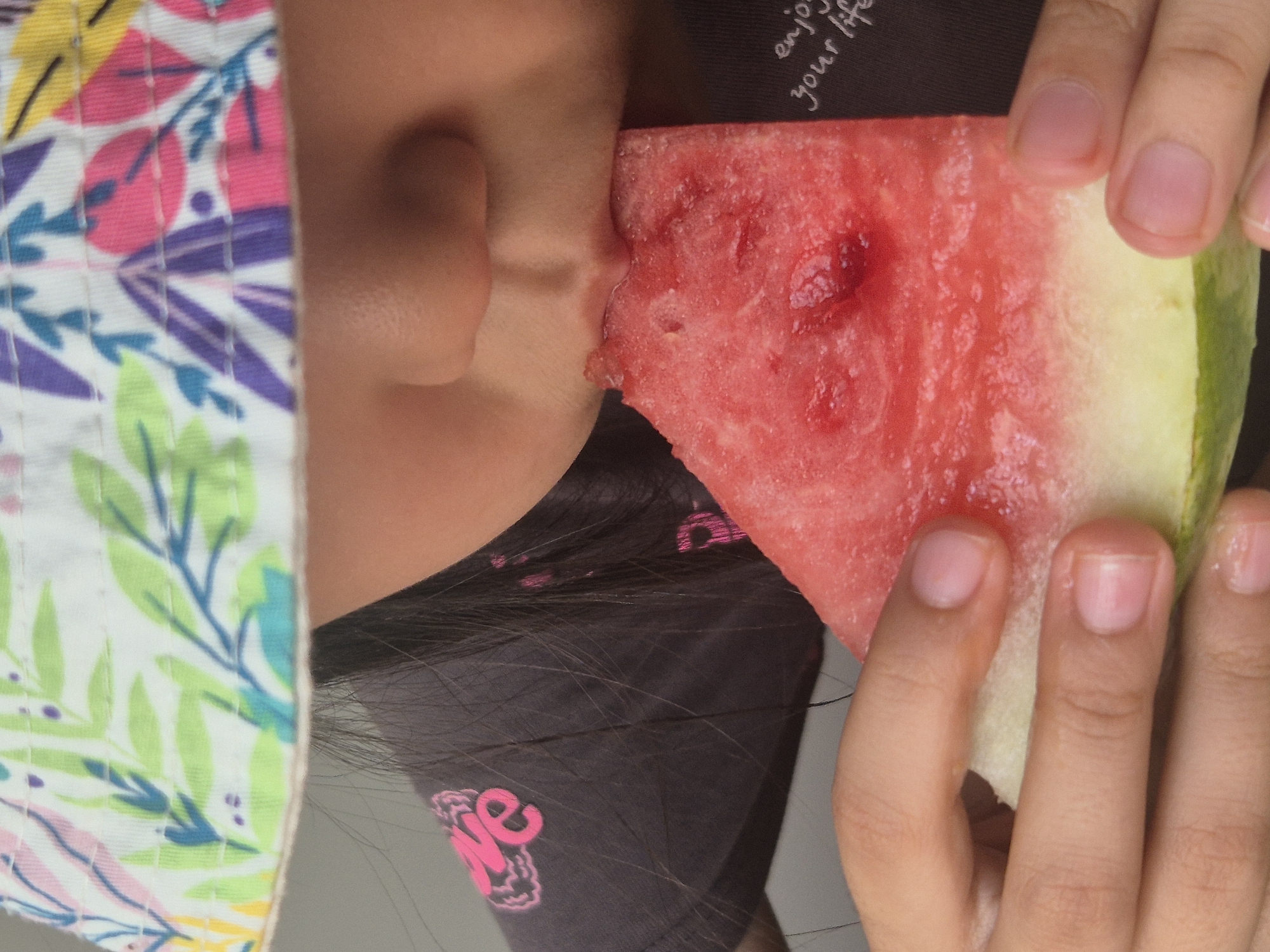 In this close-up, vertical shot, a young individual wearing a colorful, floral-patterned bucket hat is captured mid-bite into a slice of watermelon. The hat's brim partially obscures their face, revealing only a portion of their nose and mouth. Their dark hair is visible beneath the hat. The individual's upper body is clad in a dark grey t-shirt adorned with pink graphic text. The slice of watermelon is thick, displaying its characteristic pink flesh and lighter green rind, and appears juicy. The individual's hands are holding the watermelon slice, with their fingers and fingernails visible in the foreground. A faint inscription of "enjoy your life" is visible in white text on the t-shirt on the right side of the frame. The background is a neutral, grey wall. There is no identifiable information to suggest this image is located in Kokshetau, Kazakhstan.