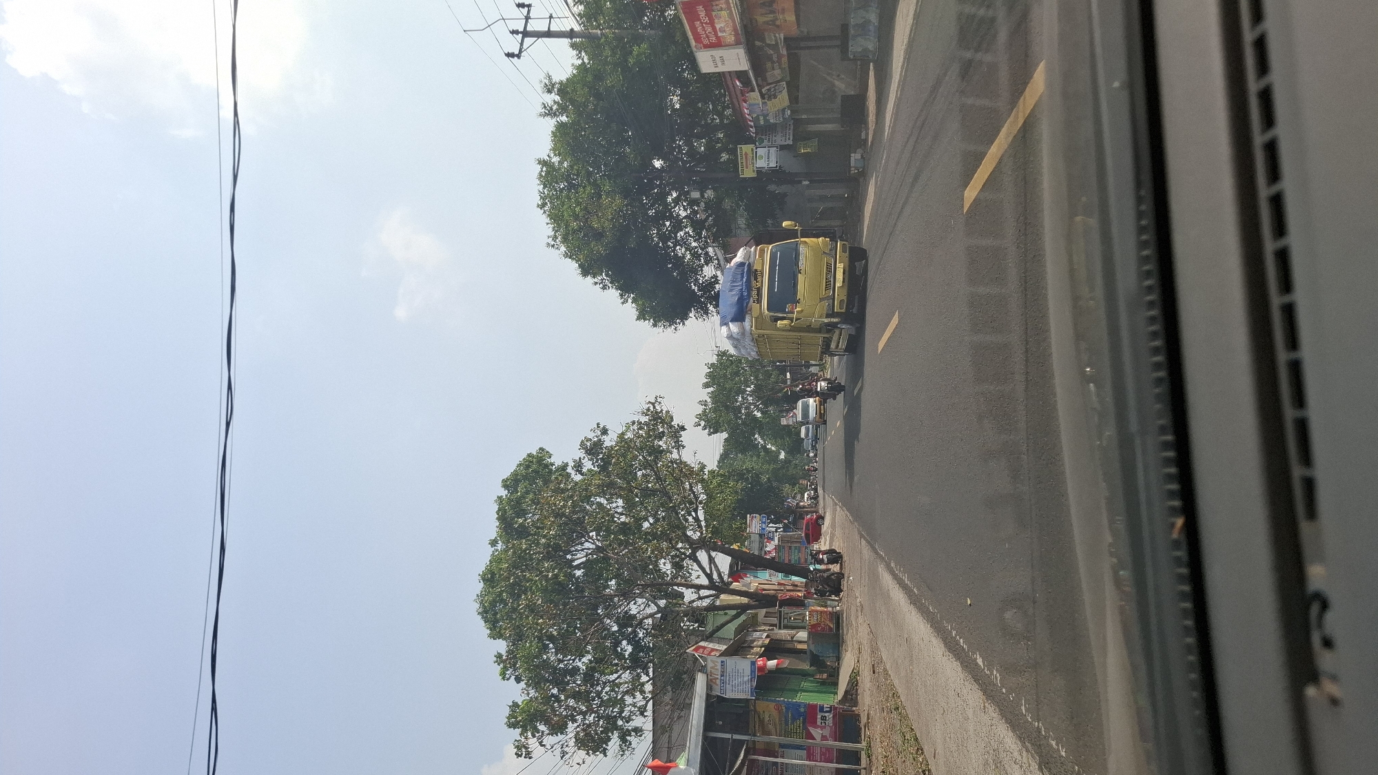 This is a photograph taken from inside a vehicle looking out at a street scene in Warungkondang, Indonesia. The perspective is from the dashboard, with the windshield in the foreground. The street is paved and appears to be a main road with two lanes of traffic. There are yellow lines marking the lanes. On the left side of the road, there are buildings with shops and signs, and several trees. A few motorcycles are parked along the sidewalk. On the right side of the road, there are also buildings and trees, with utility poles and wires visible. A yellow truck with a blue tarp covering its cargo is driving on the right lane of the road. Several other vehicles, including cars and motorcycles, are also on the road, moving in the same direction as the truck. The sky is clear and blue with some scattered clouds. The overall scene depicts a typical street view in a developed area of Warungkondang during the daytime.