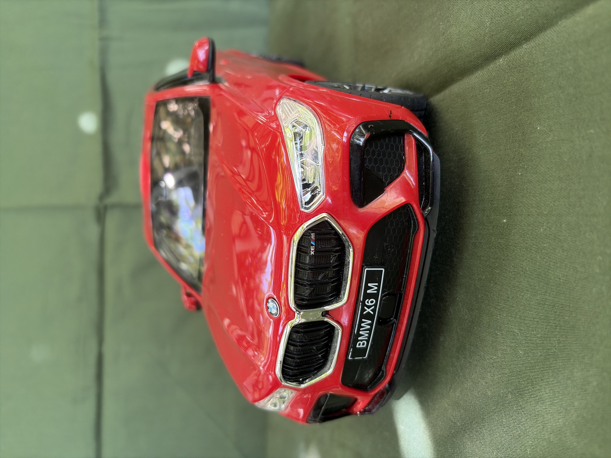 A red BMW X6 M toy car is displayed on a green textured surface. The car is facing forward, with its headlights and grille prominently visible. A license plate on the front of the car reads "BMW X6 M." The background is a blurred green surface, possibly a tablecloth or fabric. The scene is well-lit, with some light reflecting off the car's surface. There are no people or any other objects in the image. The image has been taken from a slightly low angle, looking up at the car.