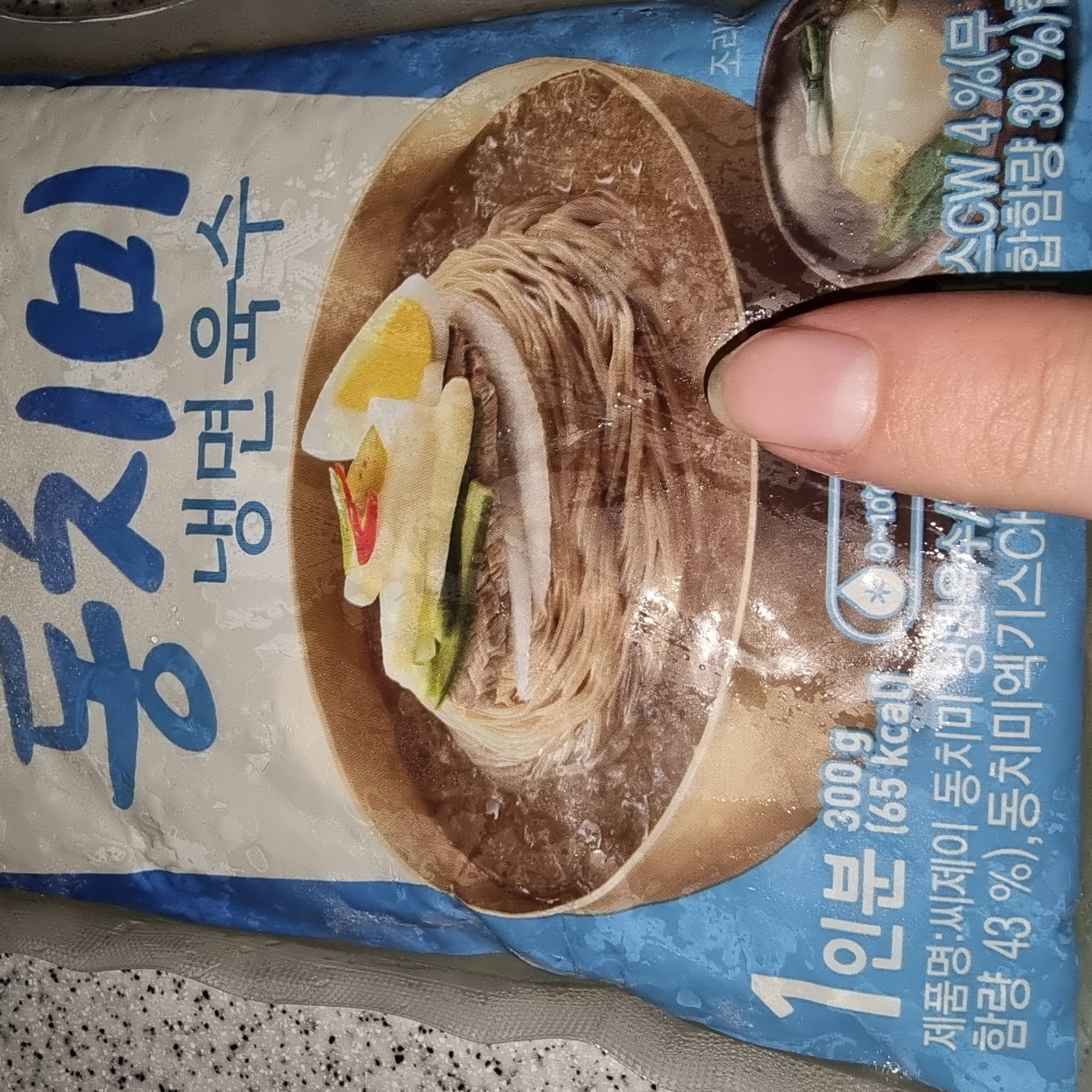 The image shows a close-up of a frozen food package of Dongchimi Naengmyeon (Korean cold noodle soup) from the brand Ssjey. The packaging features a stylized illustration of the dish, with noodles, broth, egg, and cucumber slices. Text in Korean is present on the package, including the product name, serving size (1 serving, 300g), calorie count (65 kcal), and ingredients. A human finger is visible on the right side of the image, obscuring a portion of the packaging. The background is textured and appears to be a countertop. The lighting is bright and even. There is no discernible information that can place the image in Dunnae-myeon, South Korea.