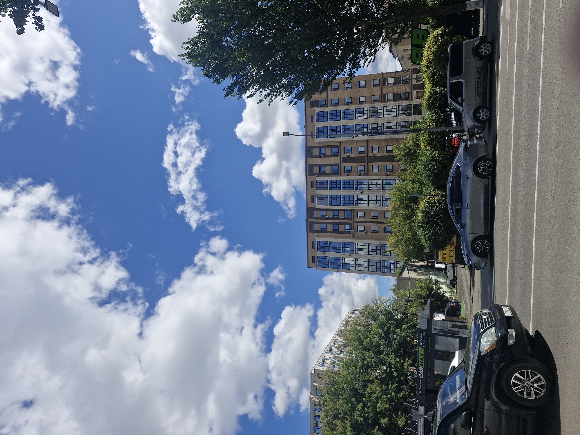 The image shows a street scene in Kokshetau, Kazakhstan, under a bright blue sky with scattered white clouds. In the foreground, a black SUV is parked on the left side of the road, facing away from the camera. Further down the road, a silver sedan and a silver minivan are also parked. Behind the parked cars, there are several modern apartment buildings with brick facades and many windows. Lush green trees and bushes are visible throughout the scene, providing a contrast to the urban architecture. The street appears to be a well-trafficked area with ongoing construction or development, as suggested by the presence of multiple buildings. The overall impression is one of a developing urban environment on a clear, sunny day.