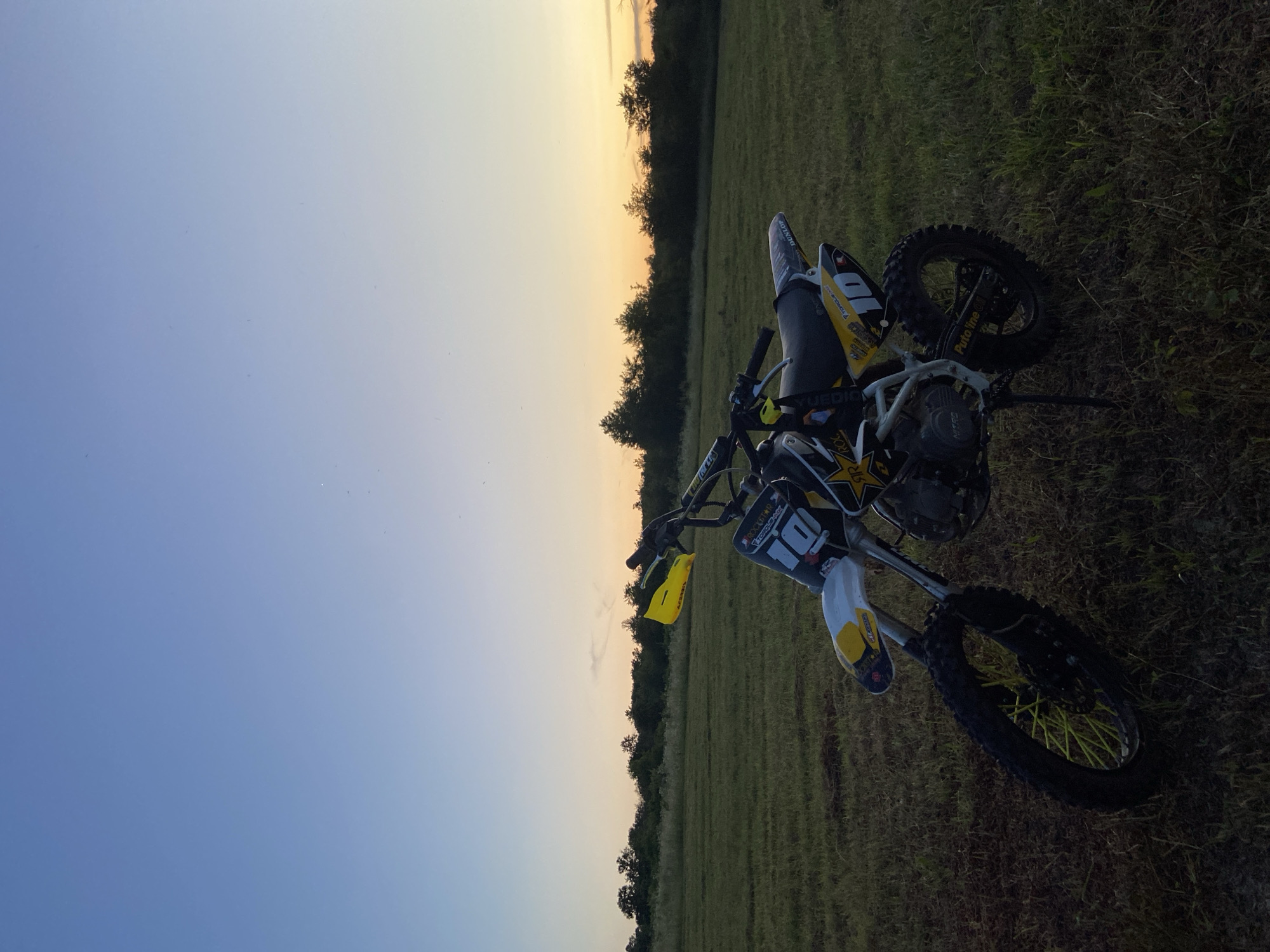 A dirt bike is parked in a grassy field during sunset. The sky is a gradient of blue to orange, with a line of dark trees in the distance. The dirt bike is black and yellow, with the number 10 prominently displayed. The front wheel has yellow spokes. The field is dry and uneven, with some patches of darker earth. The lighting suggests it is either early morning or late evening. The location is identified as Mindia, Bulgaria.