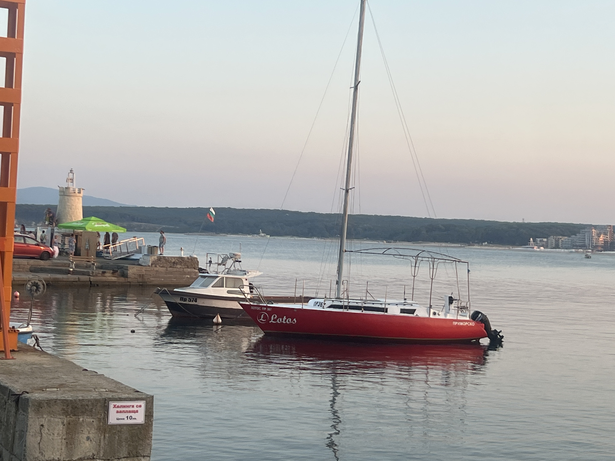 The image shows a harbor scene at dusk, likely in Primorsko, Bulgaria, given the text visible on a boat. Several boats are docked in the calm, reflective water. The most prominent is a bright red sailboat with a white superstructure and a bimini top. The name "Lotos" is clearly visible in white lettering on its side, and "ПРИМОРСКО" is written on the stern, suggesting its home port.
Behind the red sailboat, a white motorboat with the registration "Пр 574" is moored. Further back, a lighthouse with a distinctive tower stands on a rocky outcrop, accompanied by a green umbrella and what appears to be a small building or kiosk. People are visible on the dock near the lighthouse, some standing and others seated.
The sky displays a soft gradient of light blues and pinks, indicating the end of the day. The water is still, with gentle ripples reflecting the boats and the sky. A concrete wall with a sign is in the foreground, bearing Cyrillic text that reads "Хелинга се заплаща" and "Цена 10лв.", translating to "Mooring is paid" and "Price 10 BGN". The overall atmosphere is serene and peaceful, capturing a quiet moment in a coastal town.