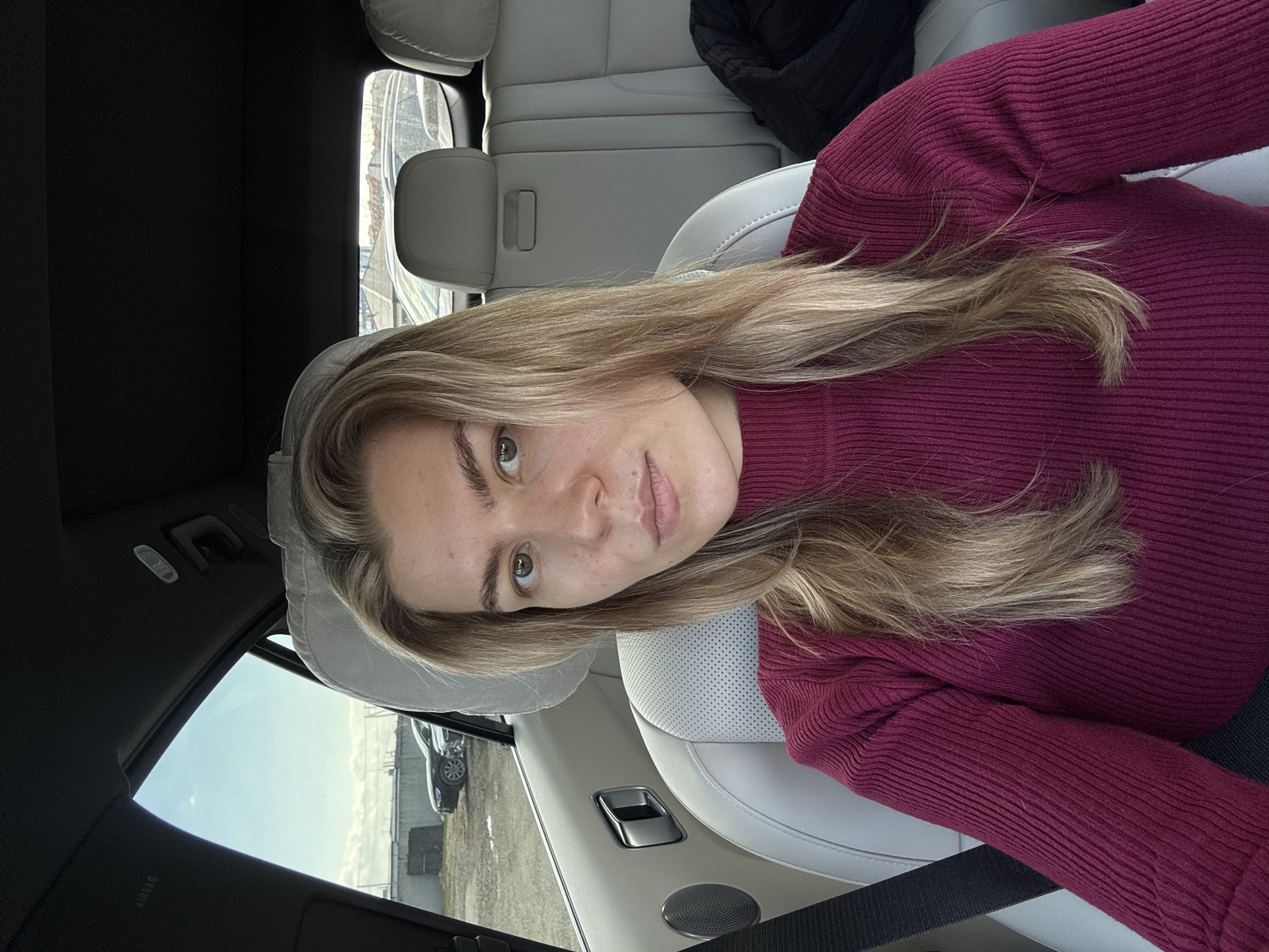 A woman is positioned in the passenger seat of a car, taking a selfie or being photographed. She has long, blonde, wavy hair and is wearing a ribbed, maroon-colored long-sleeved top. Her light-colored eyes are looking directly at the camera, and she has a neutral expression with visible freckles on her face.
The car's interior features light-colored leather seats and headrests. A black jacket or bag is visible on the back seat behind her. Through the car windows, a clear, bright sky indicates it is daytime. The background outside the car shows an open, possibly industrial or undeveloped area with some distant structures and another dark-colored car parked. A speaker grille is visible on the car door panel. The word "AIRBAG" is faintly visible on the interior panel near the driver's side window. The overall scene suggests a casual moment while traveling or parked.