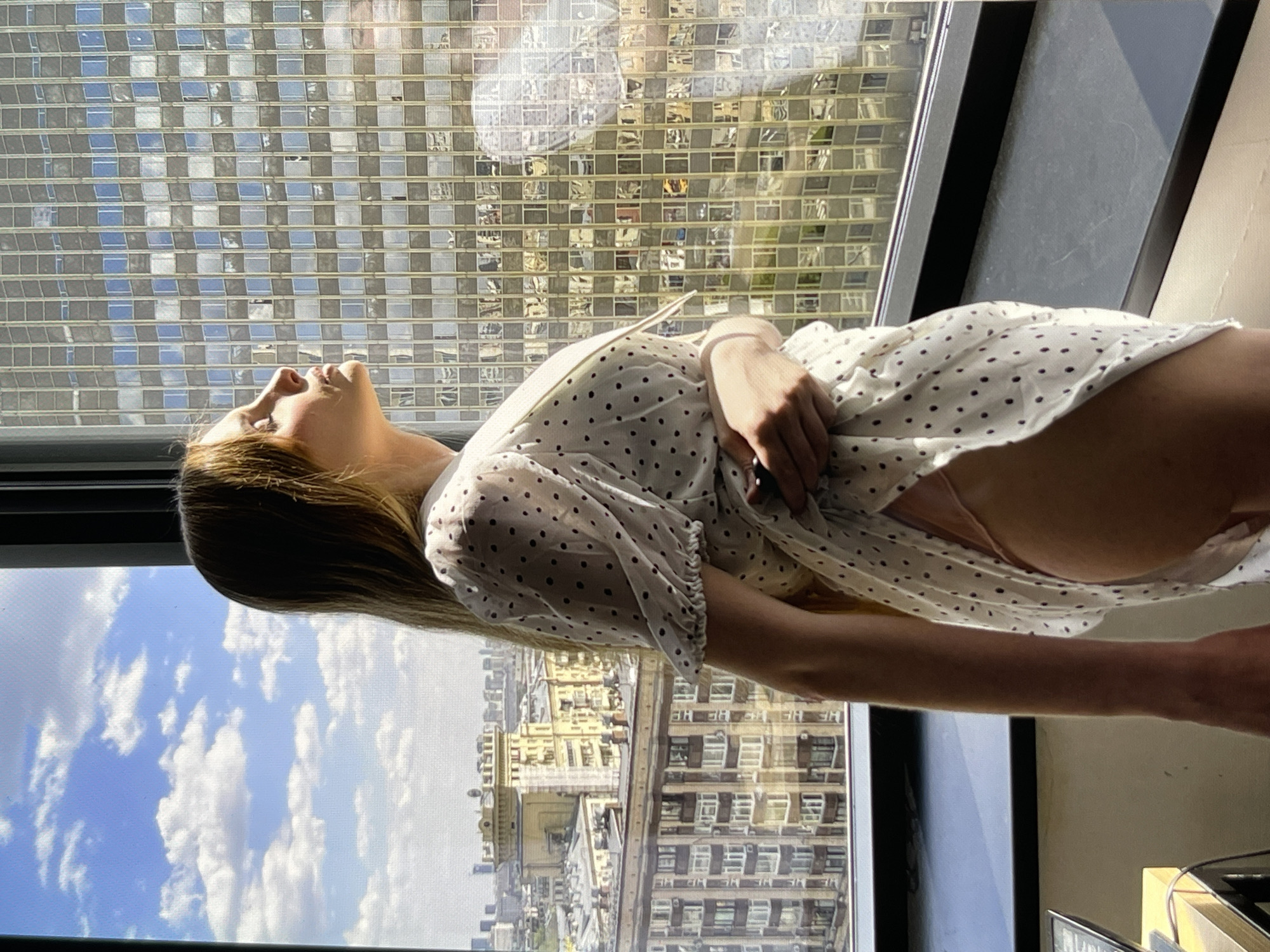 A woman with long blonde hair stands near a large window, looking out at a city skyline. She is wearing a white, short-sleeved dress with small dark polka dots and a light pink garment underneath. The window offers a view of multiple buildings, including a tall, modern structure with reflective square windows and older, more ornate buildings in the distance. The sky is blue with scattered white clouds. The woman's right arm is bent at the elbow, and her hand is near the hem of her dress, lifting it slightly to reveal her leg. Her eyes are closed, and her head is tilted back as she faces the light. The lighting suggests it is daytime, with sunlight illuminating the scene.