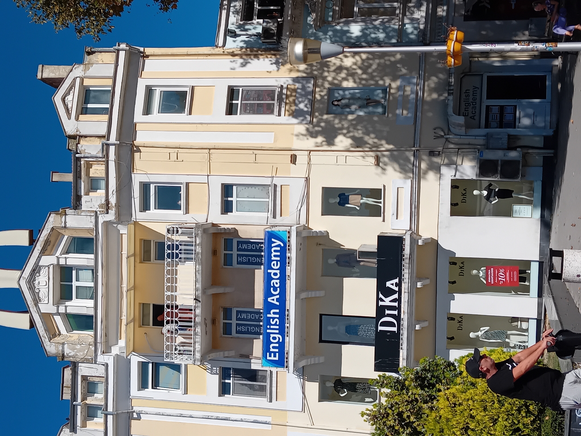 The image shows a building in Varna, Bulgaria, with several businesses and an "English Academy" on display. On the second floor, a blue banner reads "English Academy," with smaller vertical signs on either side saying "ENGLISH CADEMY." Below this, a black sign with white lettering spells out "DİKA." The building has multiple stories with windows and balconies. A man wearing a black t-shirt and a black baseball cap is standing in the foreground, leaning on a dark object. He appears to be looking to his left. In the background, a woman is sitting on a bench to the right. Trees and shadows are visible. The sky is clear and blue.