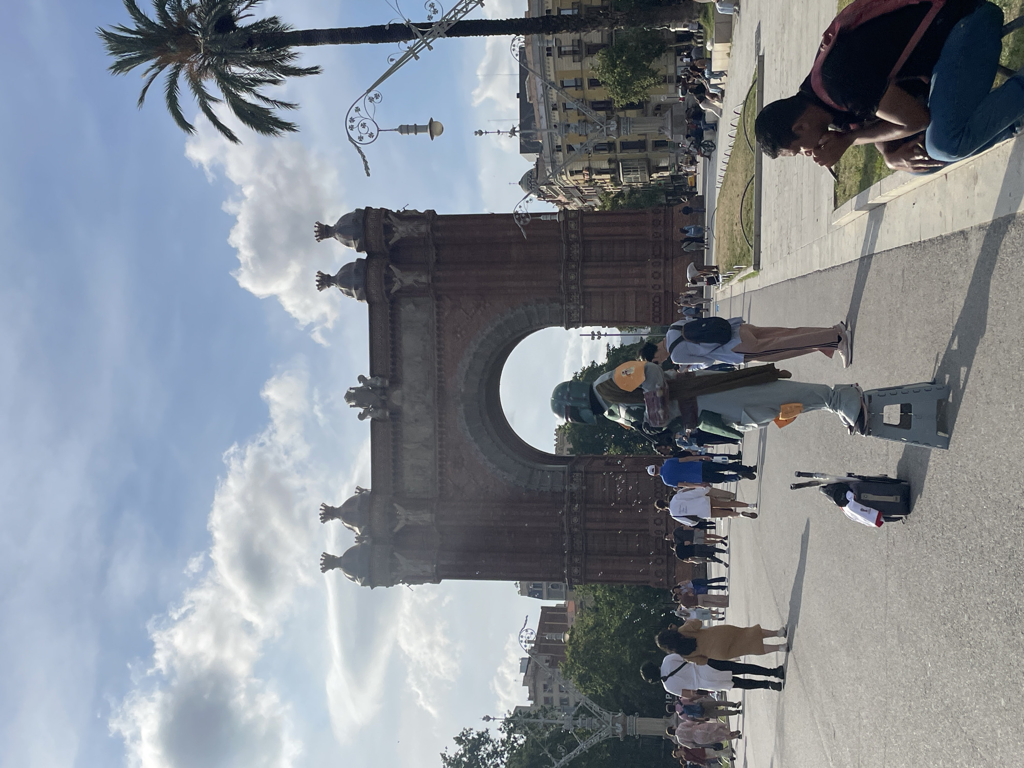This detailed image captures an outdoor urban scene in Barcelona, Spain, centered around the Arc de Triomf on a partly cloudy day.
In the foreground to the right, a male individual with dark skin, wearing a black short-sleeved shirt, blue jeans, and a red backpack, is crouched on a low concrete ledge. His right hand is positioned near his mouth. In the center, a person dressed in a Boba Fett costume (green, grey, and orange) from Star Wars stands on a grey plastic folding step stool. Small, clear bubbles are visible floating in the air around the costumed figure. To the left of the performer, on the paved ground, rests a black rolling suitcase with a white garment, possibly a t-shirt with a graphic design, draped over it. Two slender, dark objects, resembling poles, lie on the ground next to the suitcase.
The midground features numerous pedestrians traversing a wide paved plaza. Among them, a man in a white long-sleeved shirt walks alongside a woman in a light brown midi dress, both moving away from the camera. Further left, another individual wearing a light blue long-sleeved top and light-colored pants with dark stripes, carrying a black backpack, also walks away. Shadows from people and objects are distinctly cast on the paved surface, indicating direct sunlight originating from the upper left. Ornate streetlamp posts with decorative metalwork are visible on both sides of the plaza; one on the left has a multi-pronged design, while another on the right is taller and positioned next to a large palm tree.
The background is dominated by the Arc de Triomf, a grand reddish-brown brick triumphal arch. The arch features multiple decorative finials or turrets along its top edge and sculptural details. Green trees and foliage are visible to the left of the arch, through its central opening, and to its right. Beyond the arch and to its right, multi-story buildings with traditional European architecture, including balconies, are visible; one prominent building on the far right exhibits a yellow facade. The sky above is light blue with scattered white cumulus clouds.