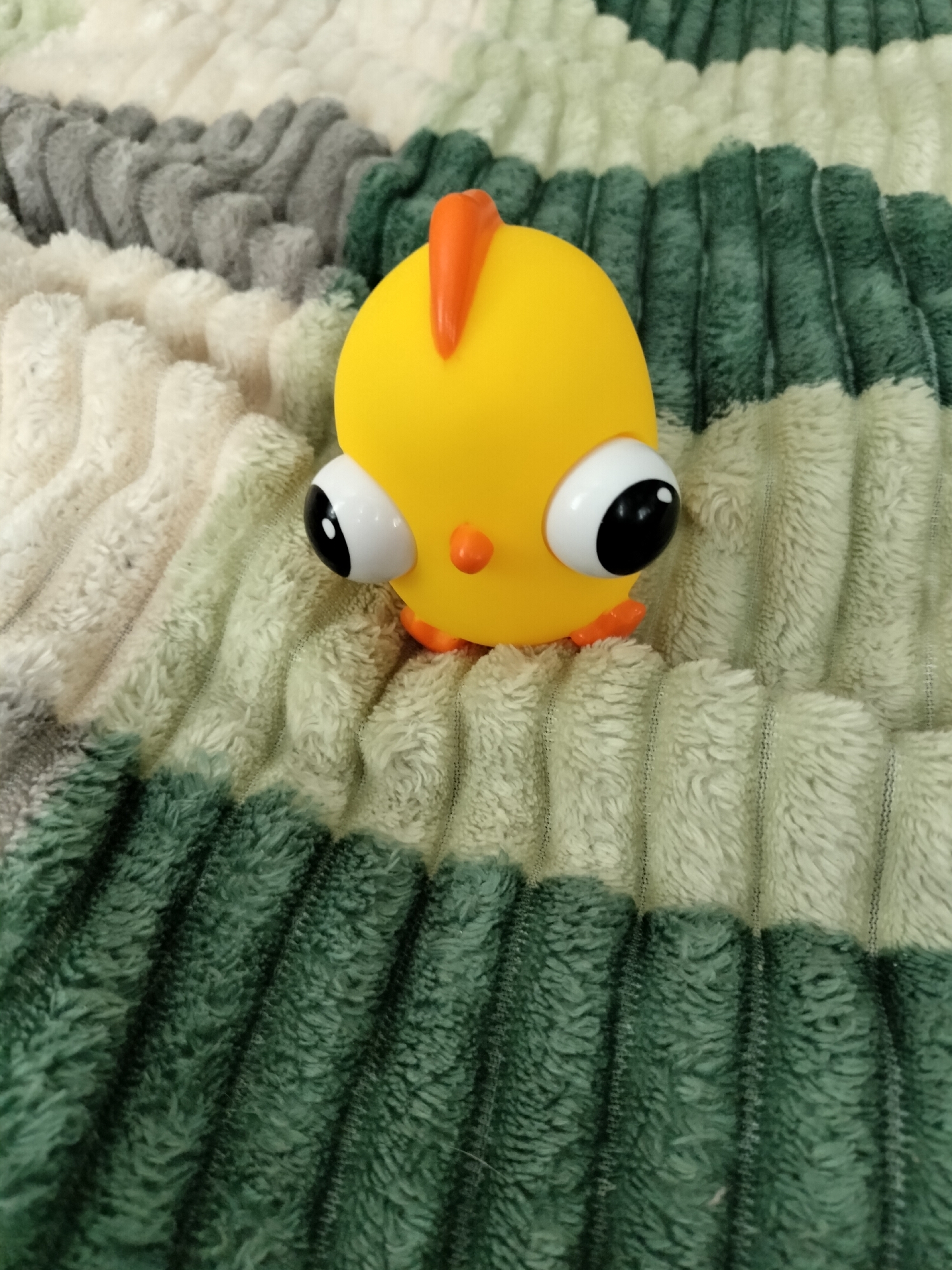 This media file is a close-up, top-down shot of a yellow rubber chicken toy. The toy has large, bulging eyes with black pupils, a small orange beak, and a single orange crest on its head. Its feet are also orange. The toy is resting on a soft, plush blanket that features horizontal ridges in alternating stripes of cream, light gray, and dark green. The texture of the blanket appears soft and slightly fuzzy. The lighting suggests an indoor setting, possibly in daylight. There is no visible text in the image. The scene is static, with the toy simply placed on the blanket. The overall impression is one of a playful and simple object.