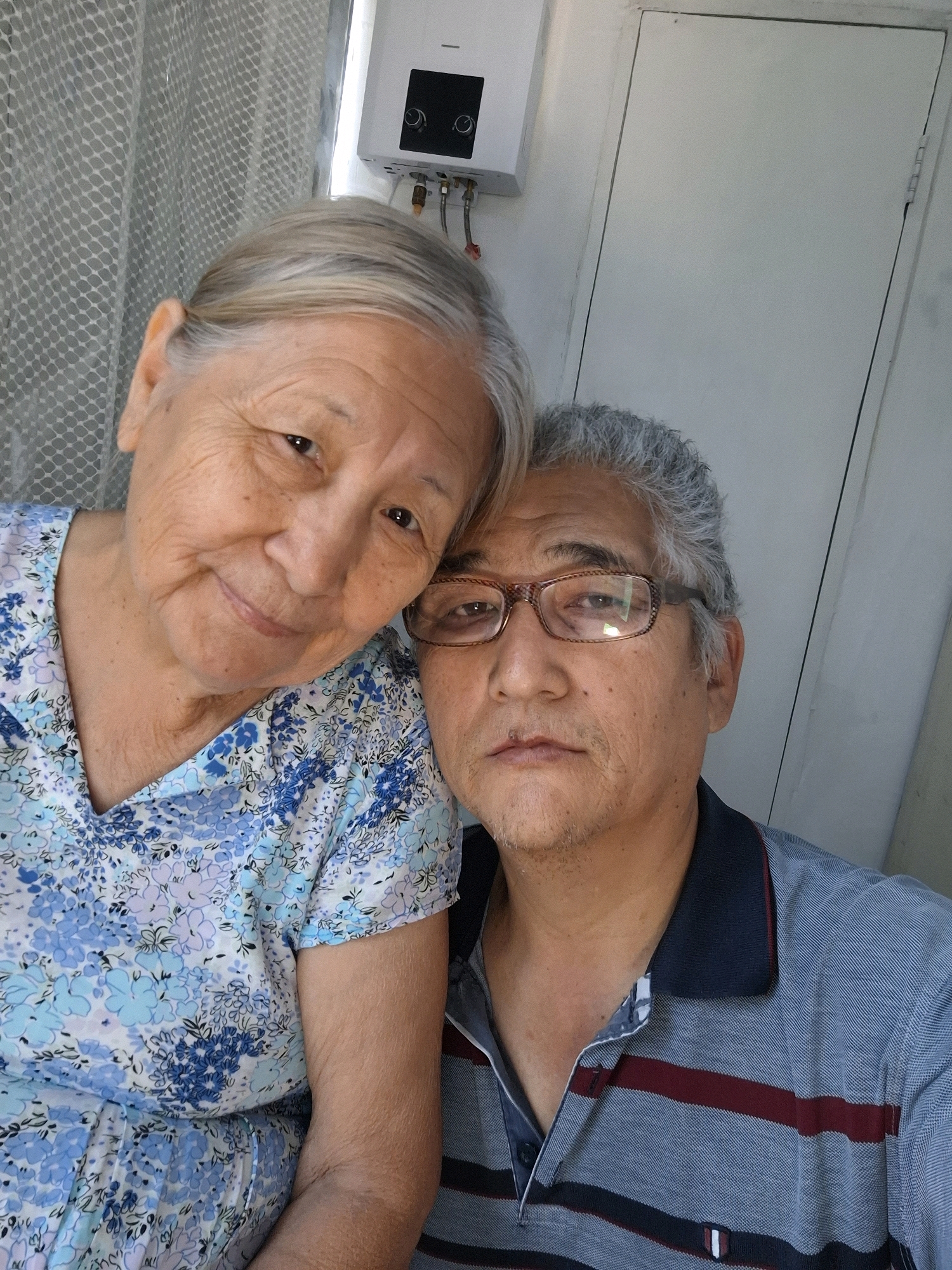 A medium shot captures two elderly individuals, an East Asian woman and an East Asian man, posing for a selfie in what appears to be an indoor setting. The woman, on the left, has graying hair and is wearing a blue floral-patterned dress. The man, on the right, has gray hair and is wearing a blue polo shirt with horizontal red stripes. He is wearing eyeglasses. Behind them, a white water heater is mounted on a pale wall, and a closed door is visible on the right. The background suggests a modest home environment, possibly in Almaty, Kazakhstan, given the context of the prompt, though there are no specific visual cues within the image to definitively confirm this location.