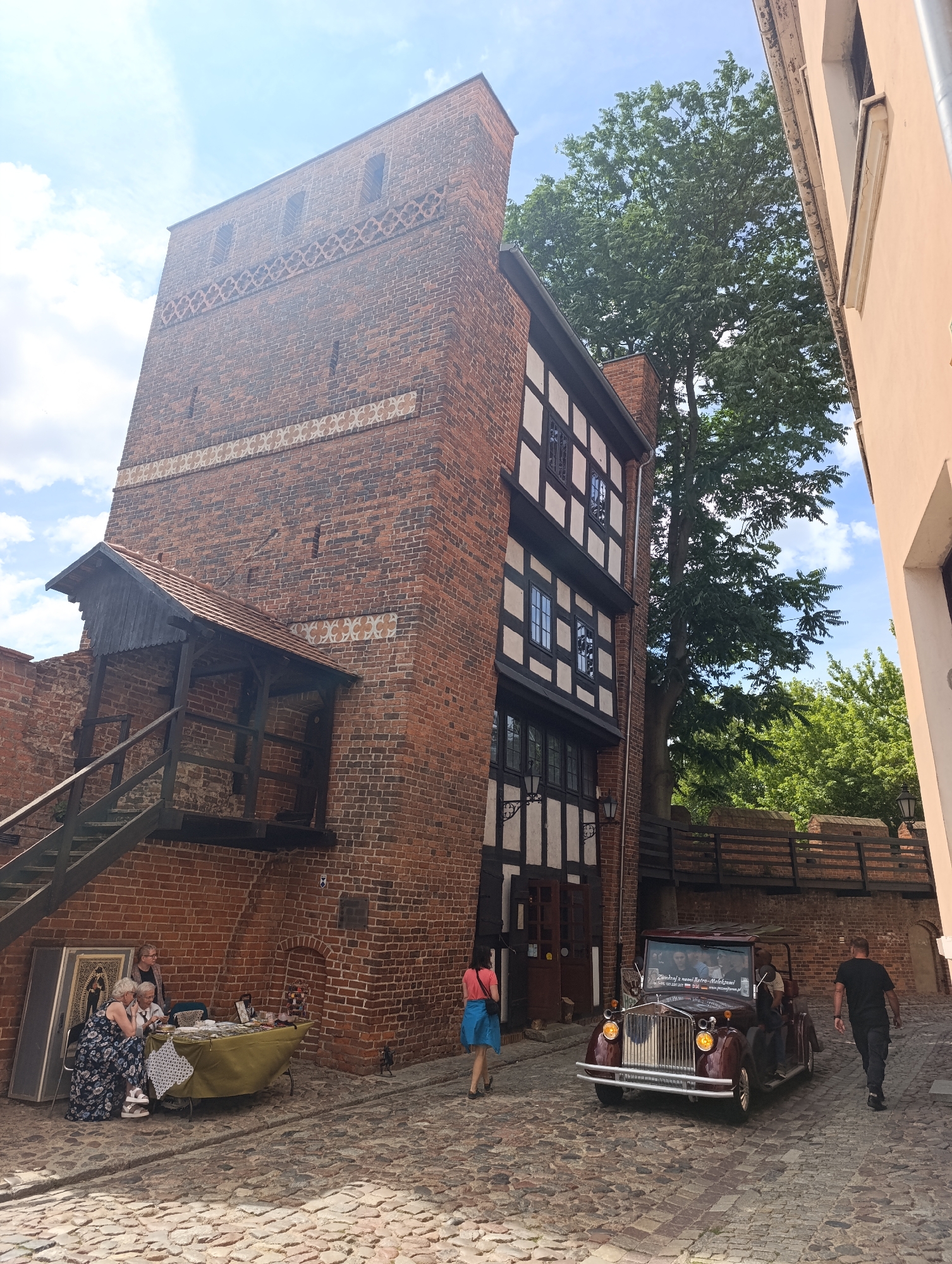 In Toruń, Poland, this image captures a cobblestone street scene dominated by a tall, red-brick tower. The tower has a unique decorative band and several small windows. Adjacent to the tower is a half-timbered building with dark beams and white infill, featuring multiple windows. An external wooden staircase with a roofed landing leads up the side of the brick tower.
On the street, a vintage, dark red convertible car is parked, carrying several passengers. To the left of the tower, three women are gathered around a table displaying various items, suggesting a small market or display. A woman in a blue skirt and pink top walks away from the car and into the distance. To the right, a man in a black t-shirt and pants walks away from the camera. Green trees are visible in the background, and the sky is partly cloudy. The architecture suggests a historical part of the city.
