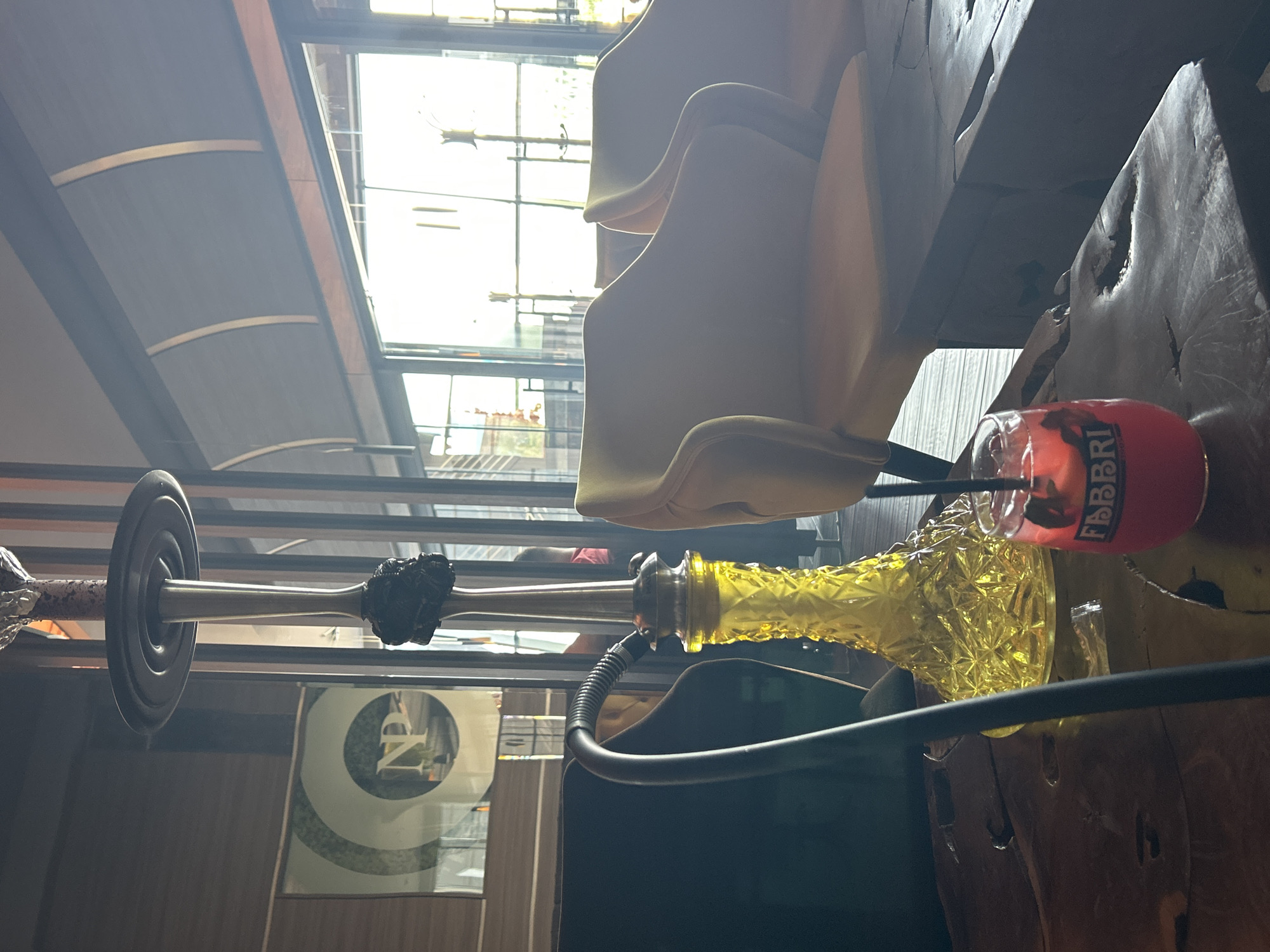The image captures an indoor scene, likely a cafe or lounge, illuminated by strong natural light. The primary subjects are a hookah and a red drink, placed on a large, irregularly textured wooden table or counter in the foreground.
A prominent hookah stands upright, featuring a decorative yellow glass base with a textured, diamond-patterned design. A dark, flexible hose extends from its base, and the metallic stem is topped with a dark bowl, possibly covered in foil. Beside it, a clear glass holds a vibrant red beverage with a straw, and the brand name "FABBRI" is clearly visible on its container or label.
In the mid-ground and background, several light-colored, possibly beige or yellow, chairs with curved backs are visible, appearing stacked or arranged in a row. They are bathed in bright natural light streaming through expansive windows or a glass wall, indicating daytime. Above, a dark, curved awning or retractable roof structure with visible supporting ribs spans the ceiling area. Through the windows, blurred elements suggest an outdoor urban environment. On a wall in the background, a framed artwork or logo displays a large, stylized letter "N" within a circular design. A dark, rectangular object, possibly a screen or monitor, is partially visible in the lower center. No people are present, but the setup suggests a relaxed and comfortable setting.