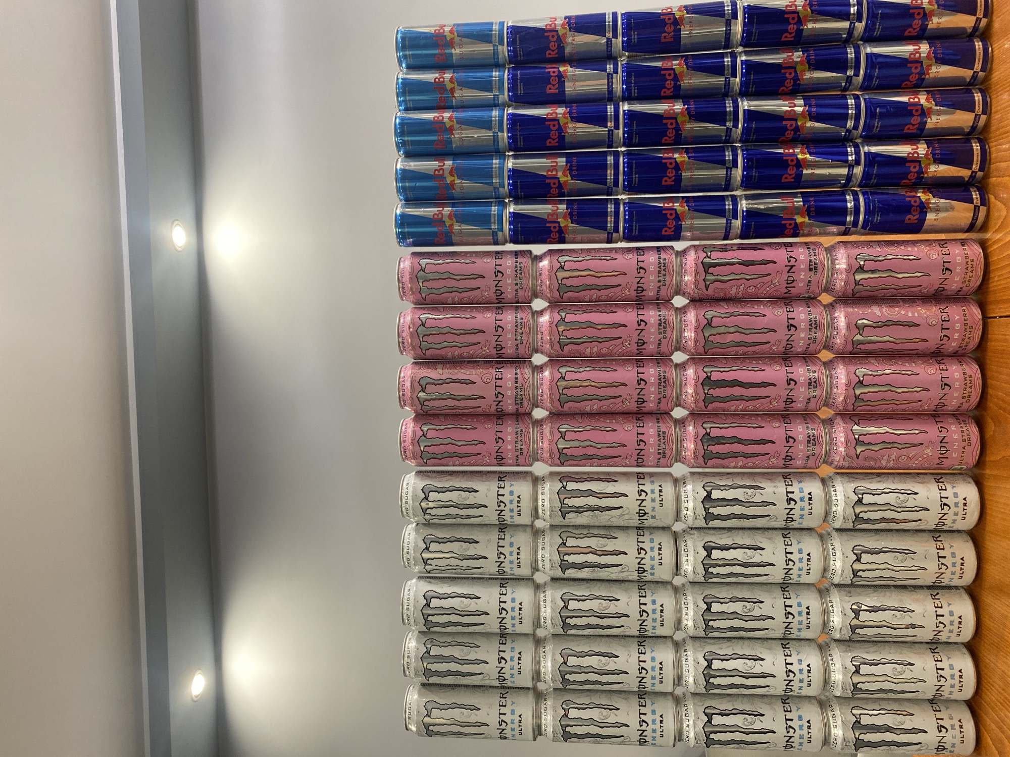 The image displays a collection of energy drink cans arranged in stacks on a wooden surface. There are three distinct brands and types of drinks visible: Monster Ultra cans (white with black logos), Monster Ultra Strawberry Dreams cans (pink with black logos), and Red Bull cans (blue and silver with red logos).
The Monster Ultra cans are arranged in four stacks, with each stack containing three cans. The Monster Ultra Strawberry Dreams cans are arranged in two stacks, also with three cans per stack. The Red Bull cans are arranged in three stacks, each containing three cans.
The scene appears to be indoors, with a grey wall and ceiling visible in the background. There are spotlights illuminating the area from above. The overall lighting suggests a well-lit environment, possibly a store display or a personal collection. There are no people visible in the image.
The cans are primarily positioned in a neat, organized manner, indicating they are likely for sale or display. The arrangement of the cans suggests a tiered display, with the front row of cans being the most visible. The reflection of the cans on the wooden surface adds a subtle visual depth to the image.